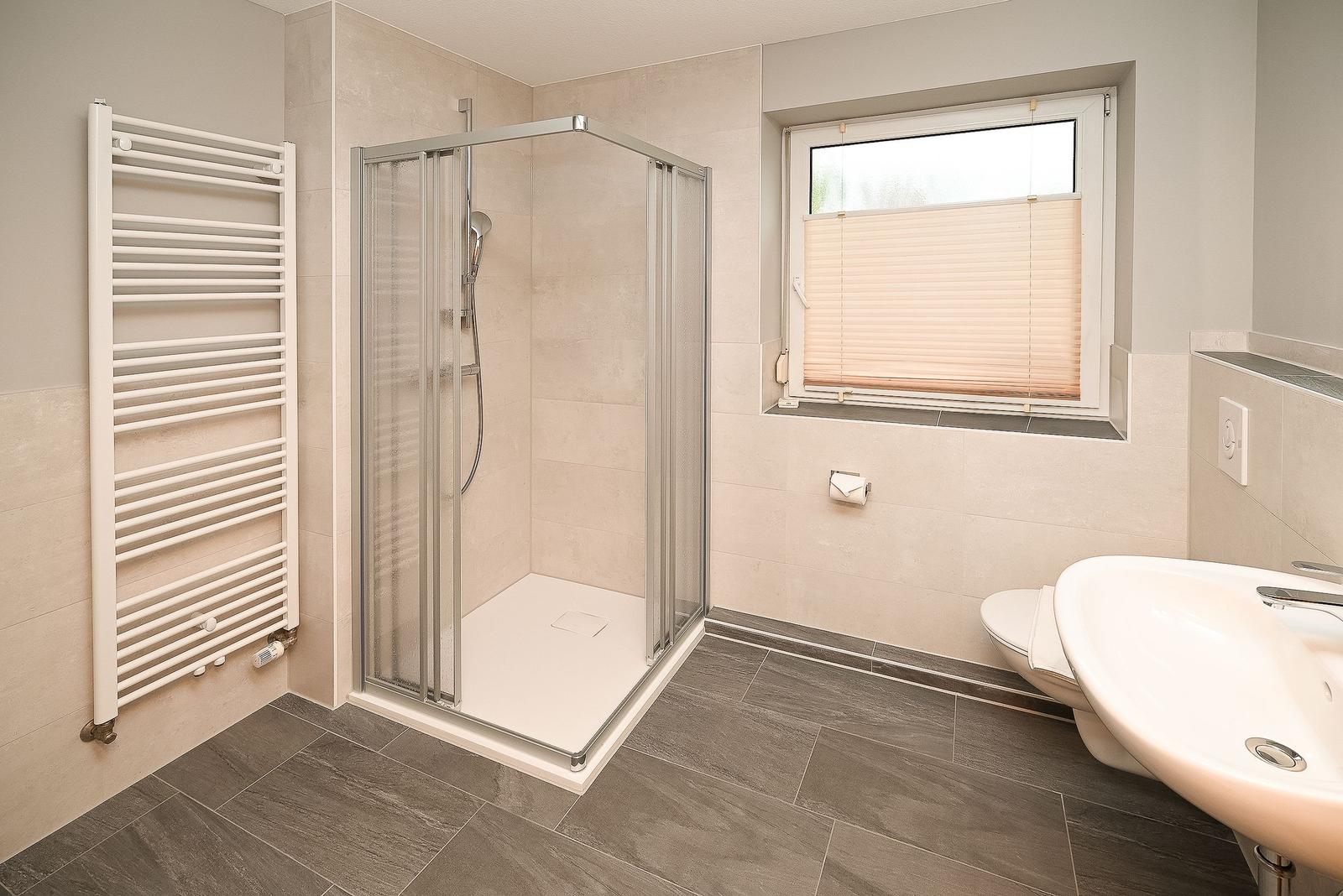 Badezimmer mit Dusche, Toilette und Waschbecken. Fenster mit Jalousie und Heizkörper.