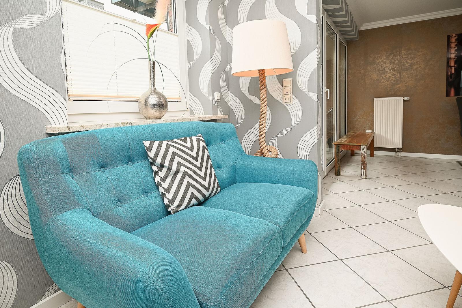 Ein blauer Sessel mit Kissen steht vor einer Wand mit Muster. Eine Lampe und ein Fenster sind sichtbar.