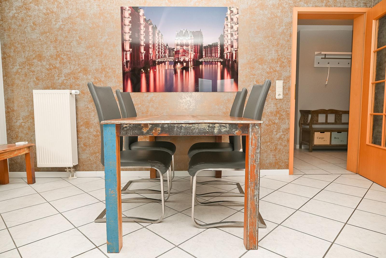 Esstisch mit vier Stühlen vor Wandbild. Tür führt in Schlafzimmer.