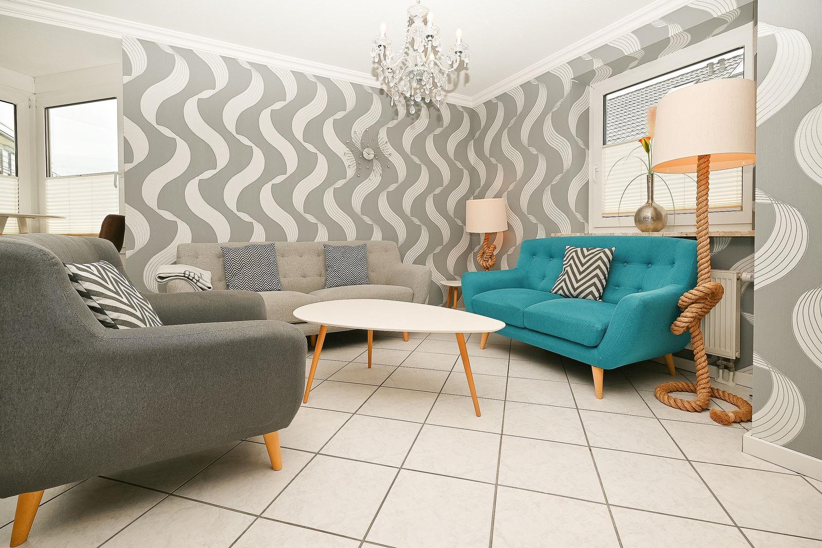 Wohnzimmer mit grauem Sofa, türkisfarbenem Sessel und Wellenförmigem Tapetenmuster an den Wänden.
