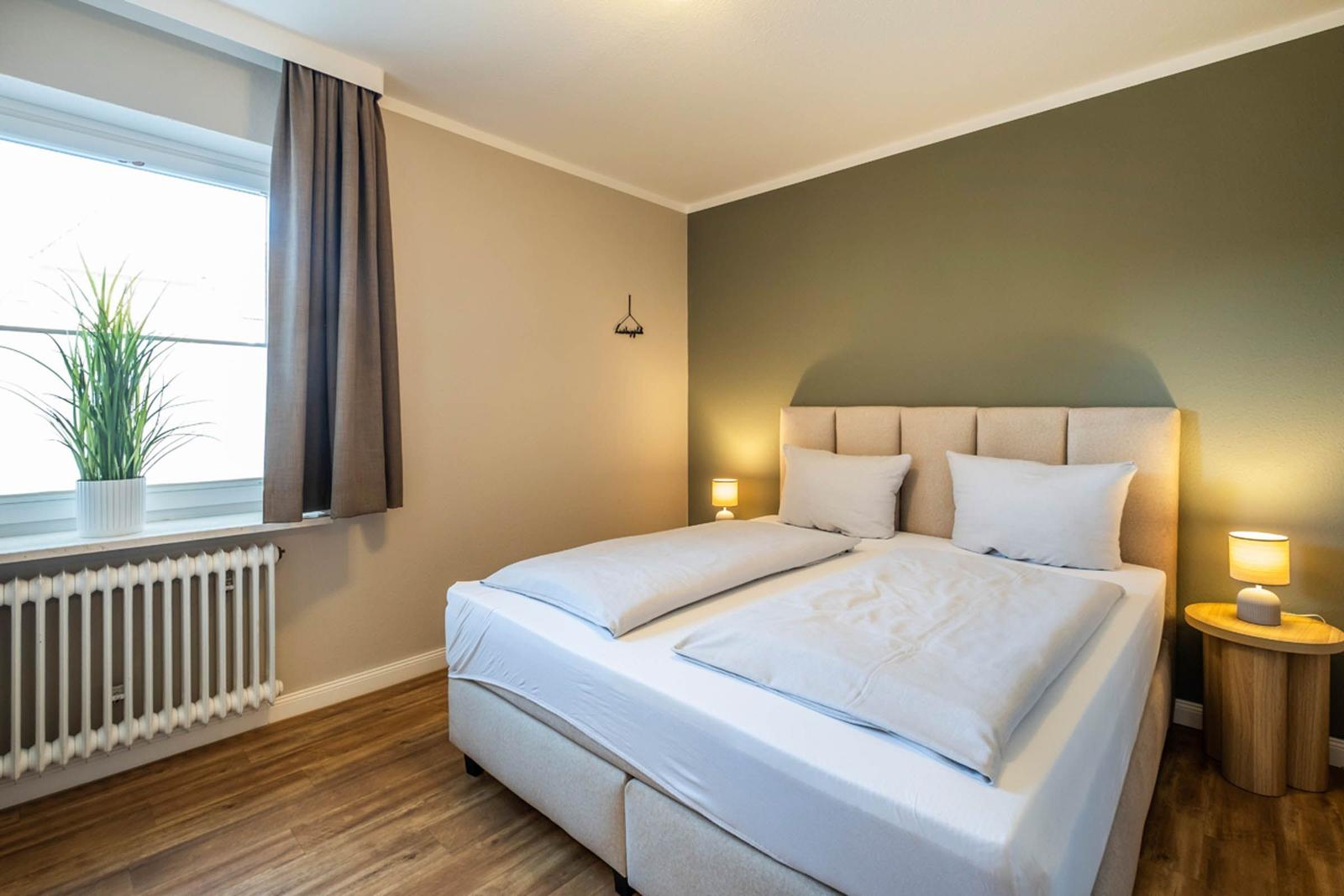 Schlafzimmer mit Bett, Nachttisch, Lampe und Fenster mit Pflanze.