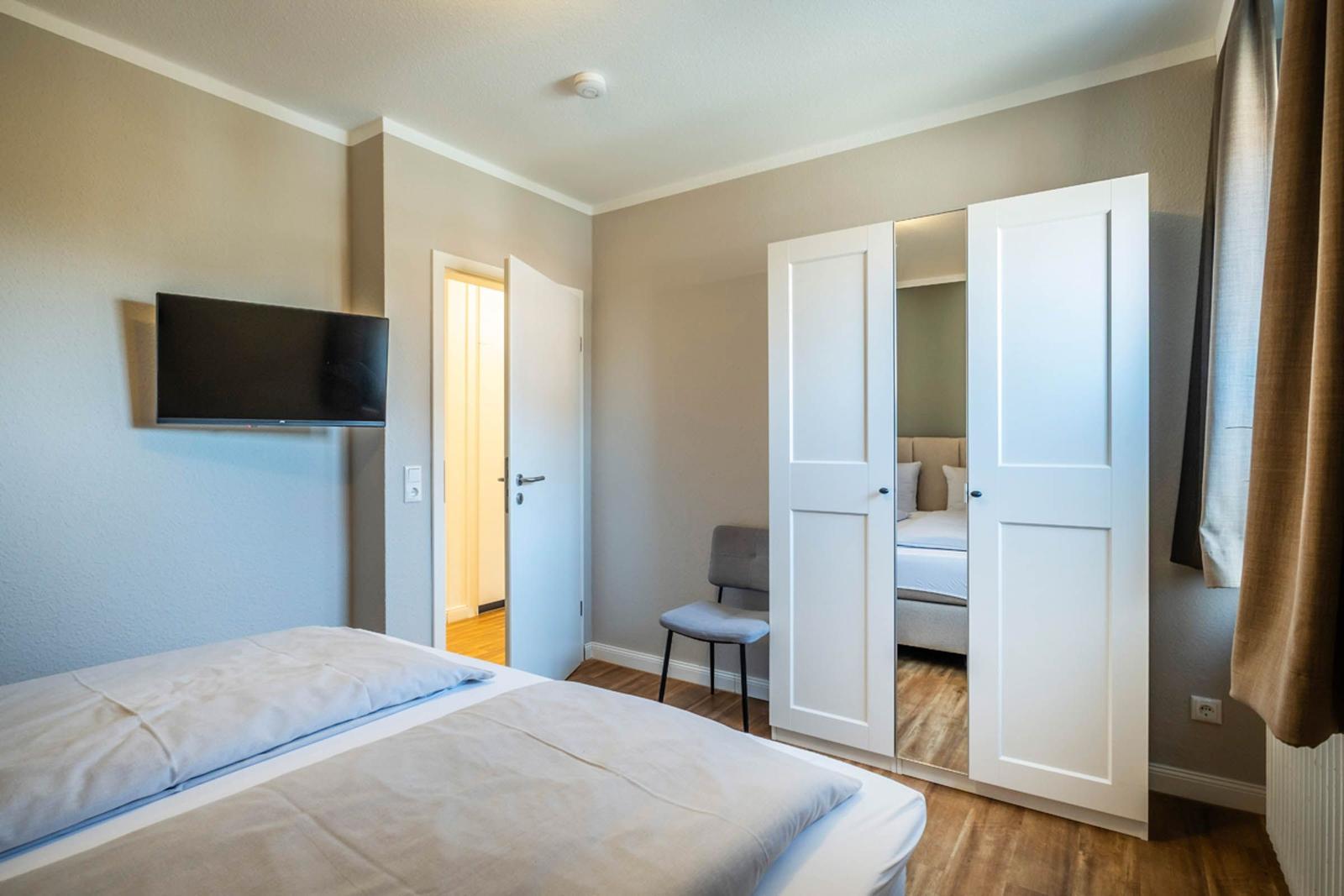 Schlafzimmer mit Bett, TV und Kleiderschrank. Tür führt in einen Flur.