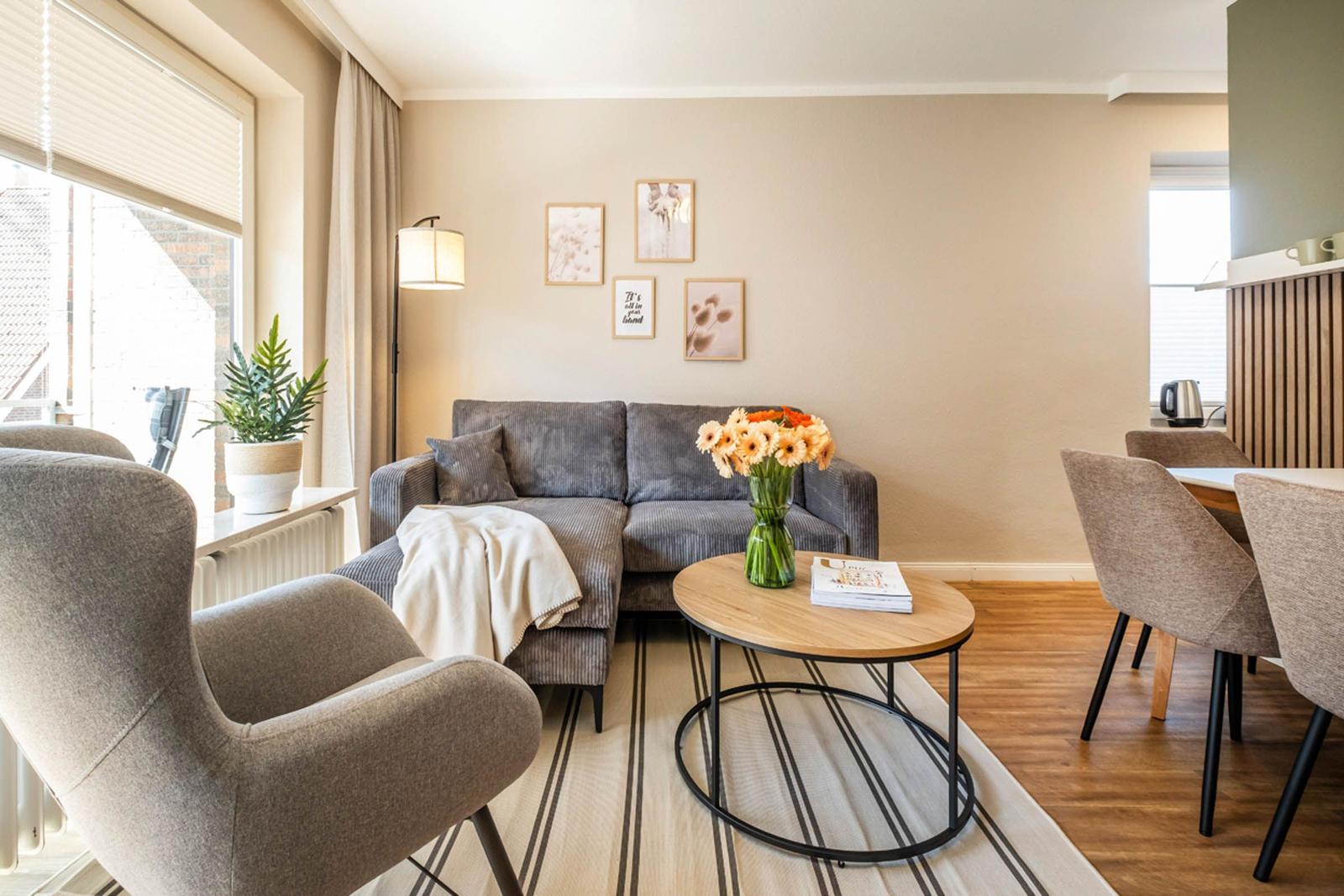 Wohnbereich mit Sofa, Sessel und Esstisch. Fenster, Wandbilder und Blumen sind sichtbar.