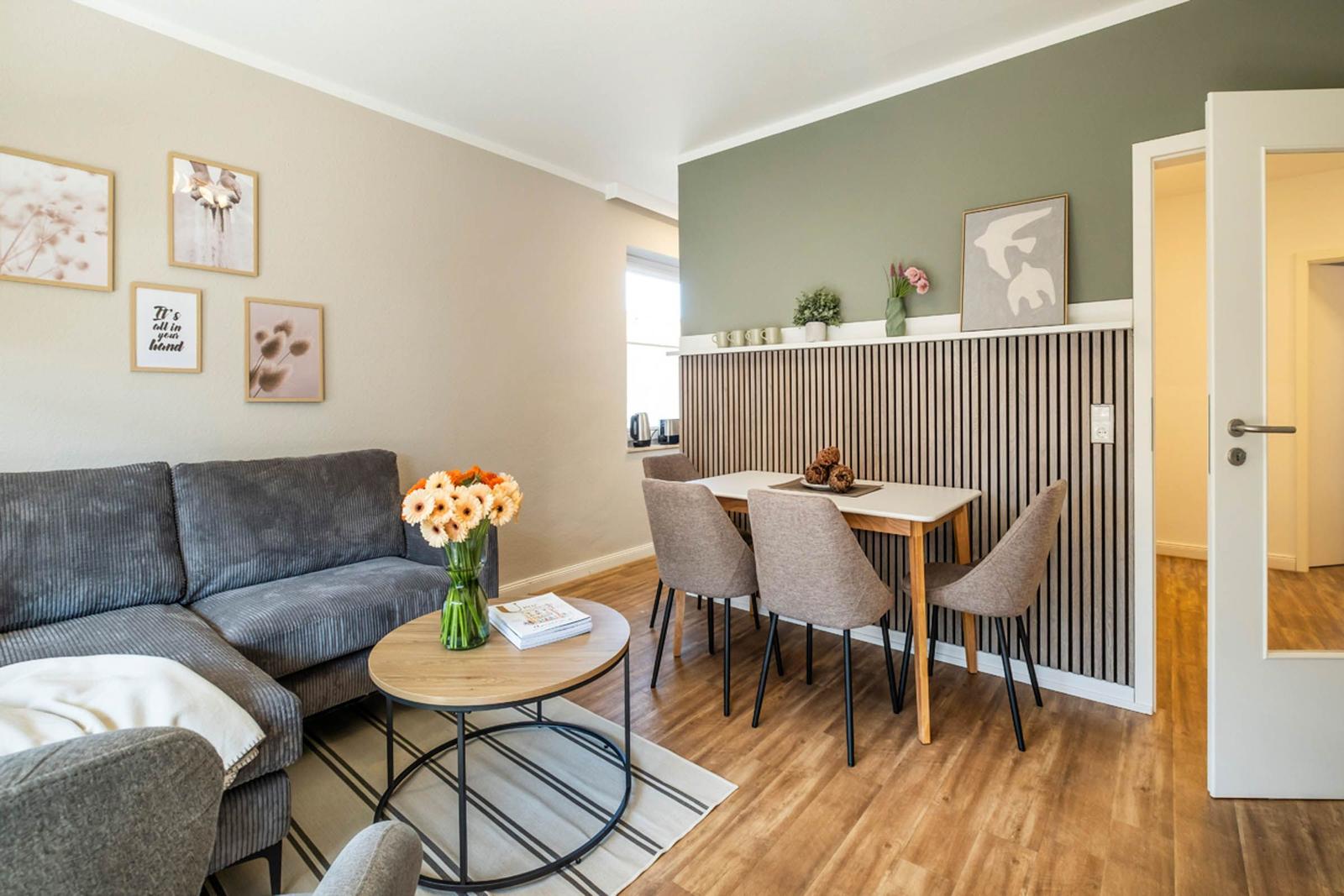 Wohn- und Essbereich mit Sofa, Tisch und Stühlen. Wandgestaltung mit Bildern und Holzpaneelen.