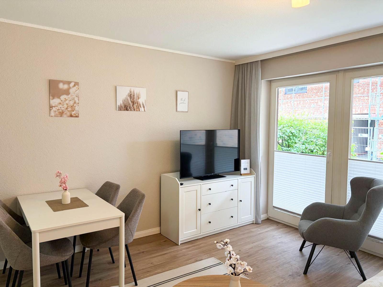 Living and dining area with TV, table, chairs, and armchair. Window with view of balcony.