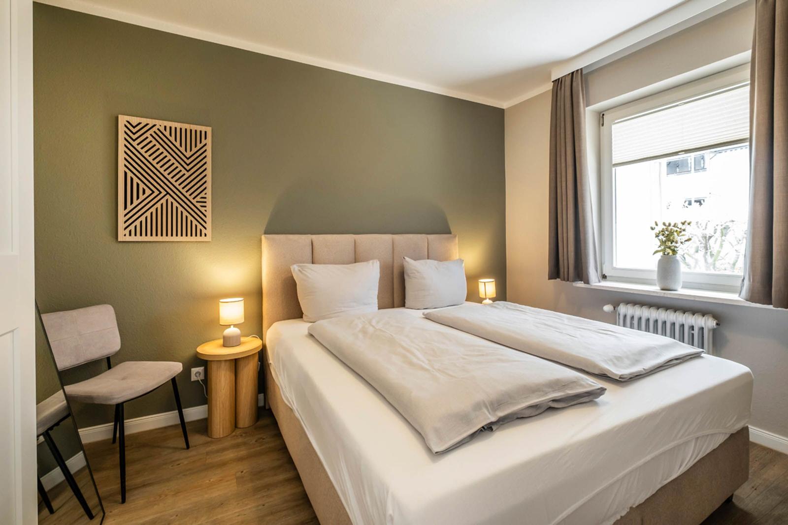 Ein Schlafzimmer mit Doppelbett, Nachttisch, Stuhl und Fenster mit Vorhang.