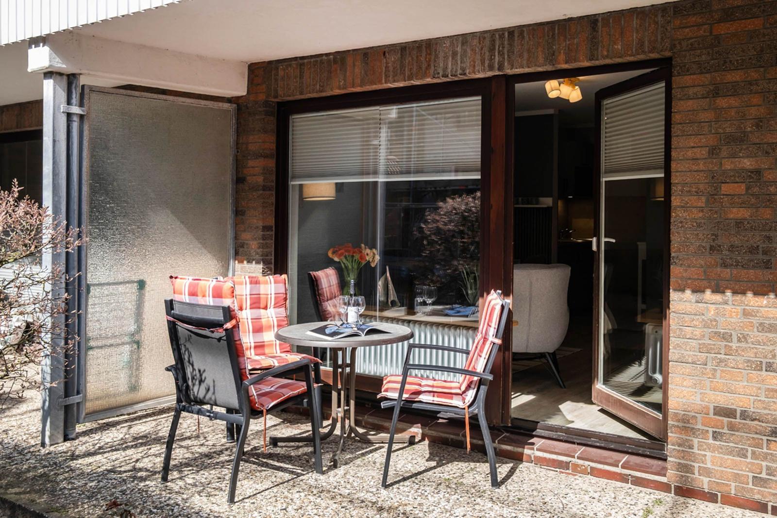 Terrasse mit Tisch und zwei Stühlen mit karierten Sitzkissen. Durch Glastür ist Wohnbereich sichtbar.