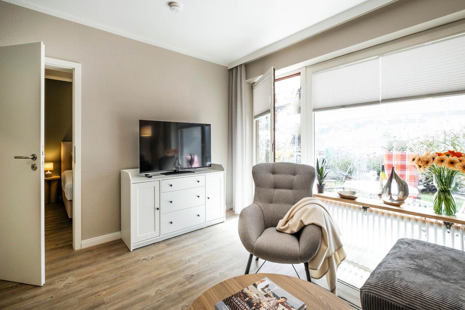 Wohnzimmer mit Fernseher, Sessel und Fensterblick. Tür führt ins Schlafzimmer.