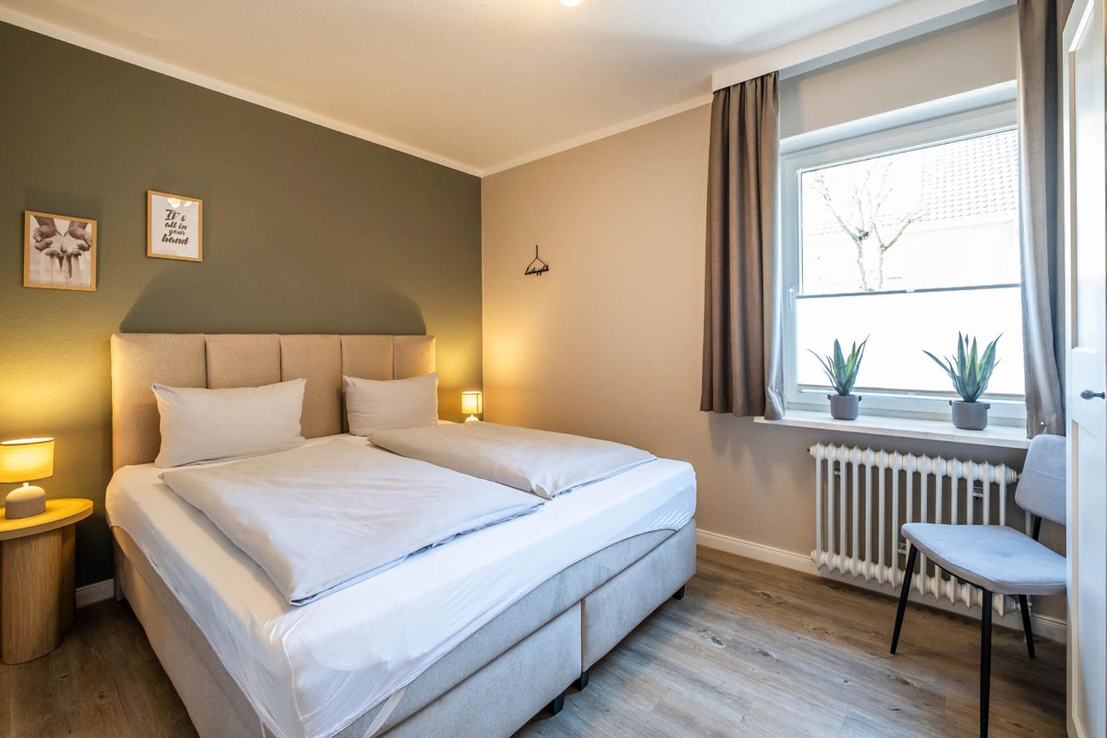 Ein Schlafzimmer mit Doppelbett, Fenster, zwei Pflanzen und einem Stuhl.