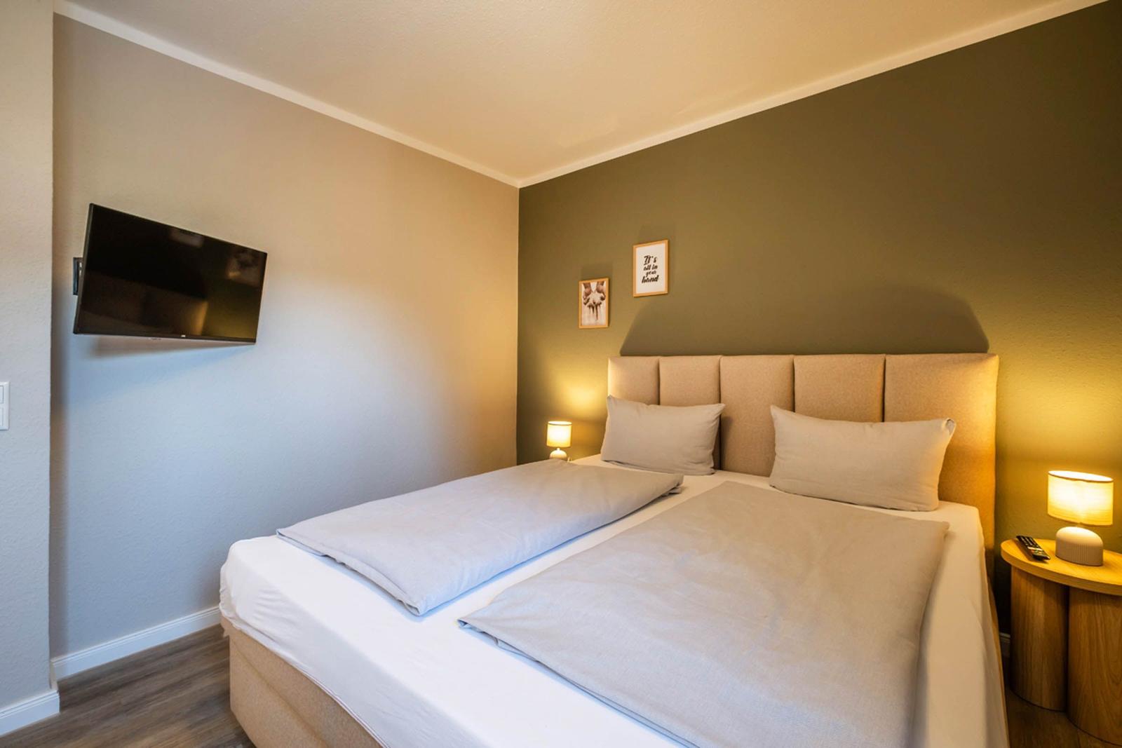 Schlafzimmer mit Doppelbett, Wandfernseher und Nachttisch mit Lampe.