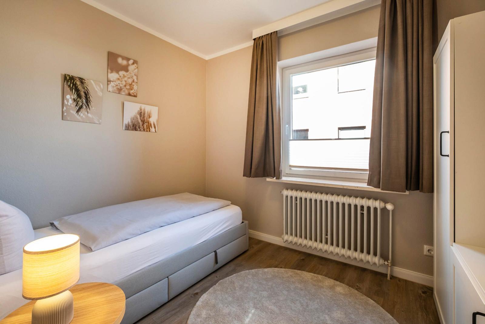Ein Schlafzimmer mit Bett, Nachttisch, Lampen, Wandbildern, Fenster und Vorhängen.