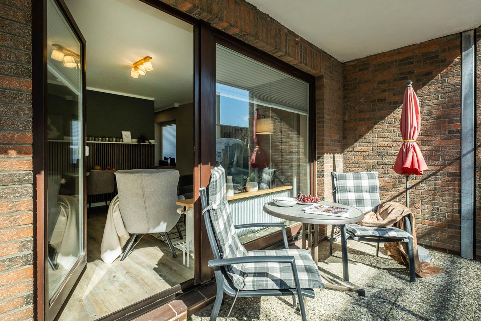 Terrasse mit Tisch, zwei Stühlen und geschlossenem Sonnenschirm. Blick ins Innere mit Sitzgelegenheit.