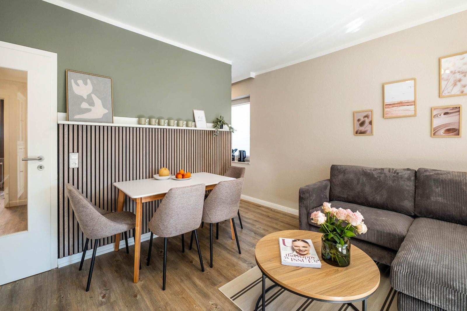 Wohn- und Essbereich mit Sofa, Tisch und Stühlen. Wand mit Holzvertäfelung und Bildern.
