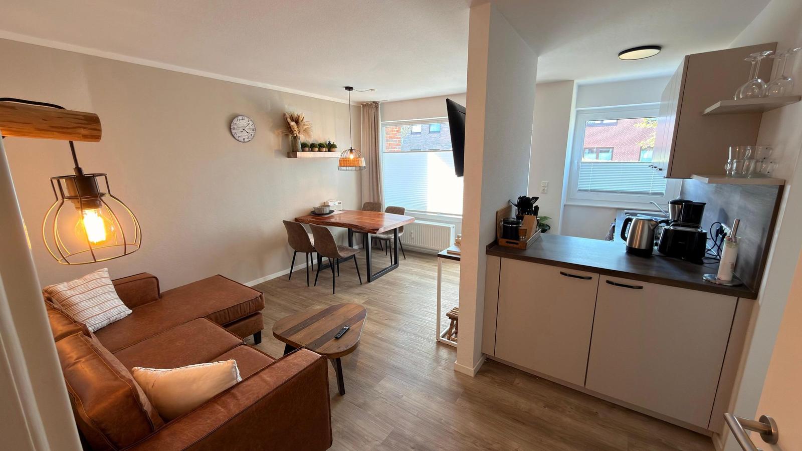 Wohnzimmer mit Sofa, Esstisch und Küche. Holzboden und Fenster mit Jalousien.