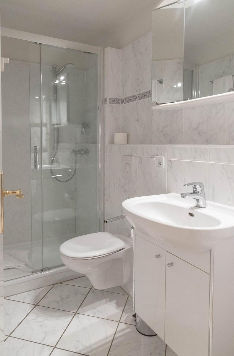 Badezimmer mit Dusche, Waschbecken und WC. Marmorfliesen und Spiegelschrank.