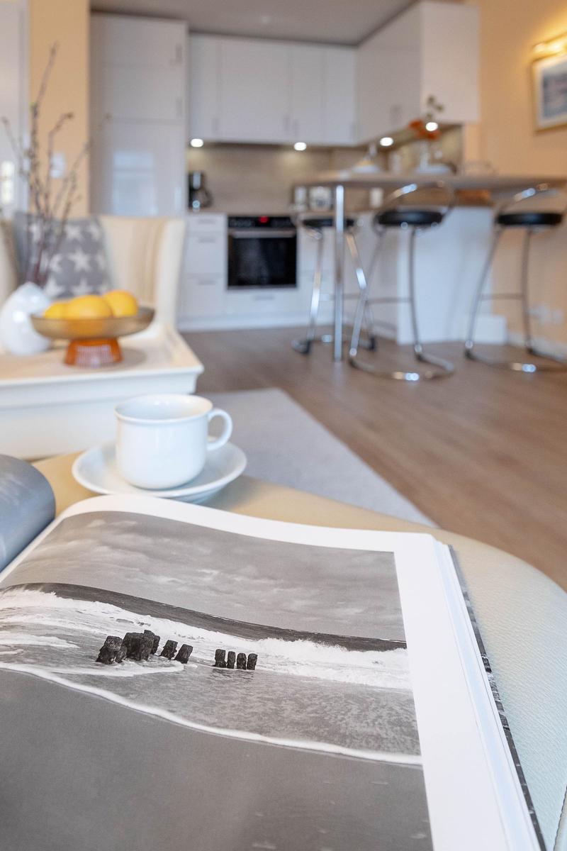 Auf dem Sofa liegt ein Buch mit Strandbild. Im Hintergrund Küche mit Barhocker.