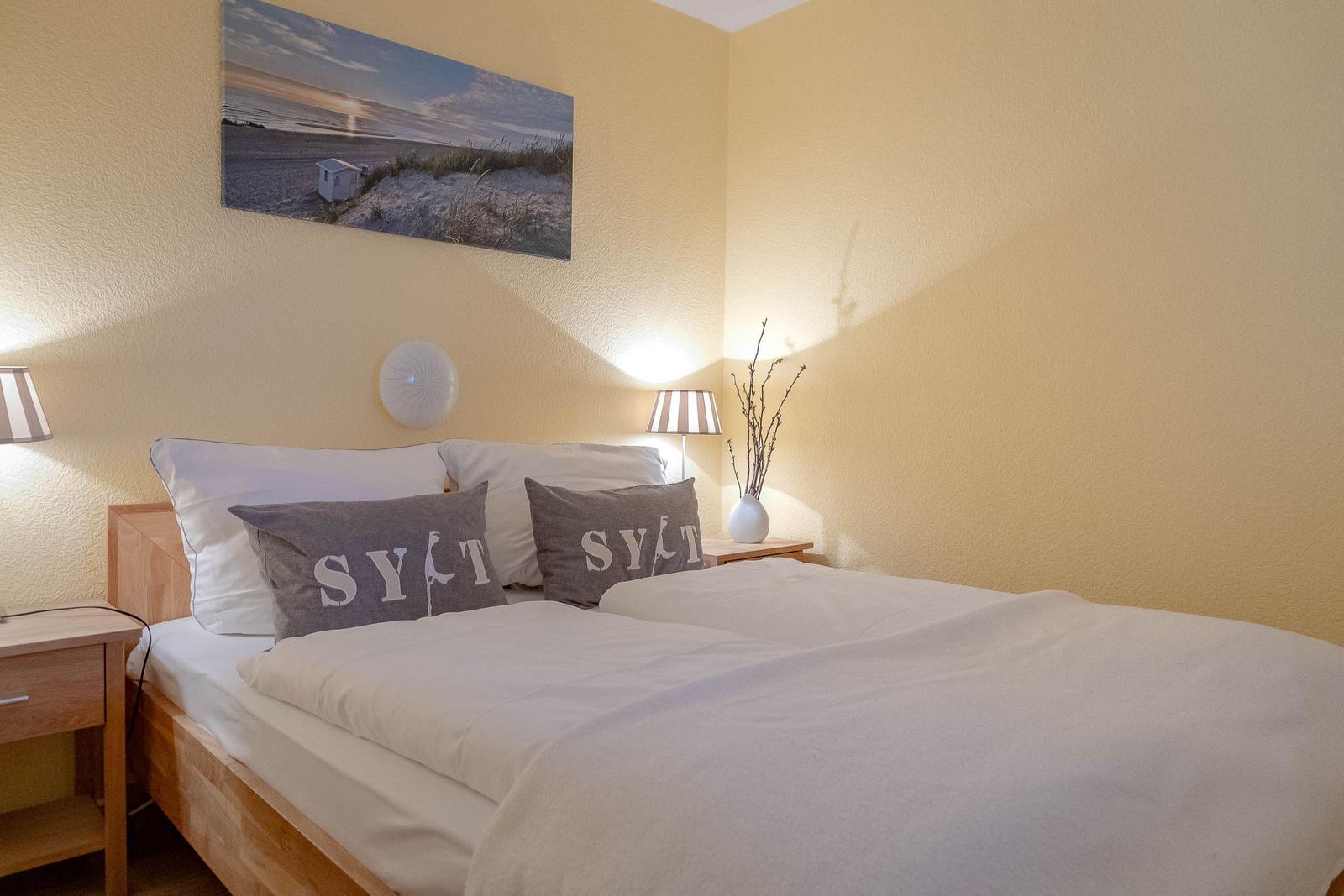 Schlafzimmer mit Bett, zwei Nachttischen und Wandbild. Kissen zeigen SYLT.