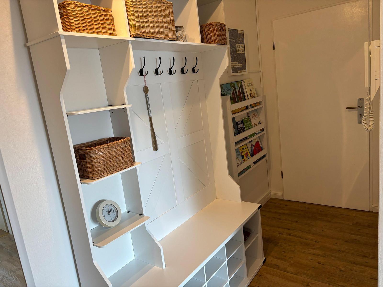Eingangsbereich mit weißen Regalen, Haken und Baskets. Holzboden und Tür im Hintergrund.
