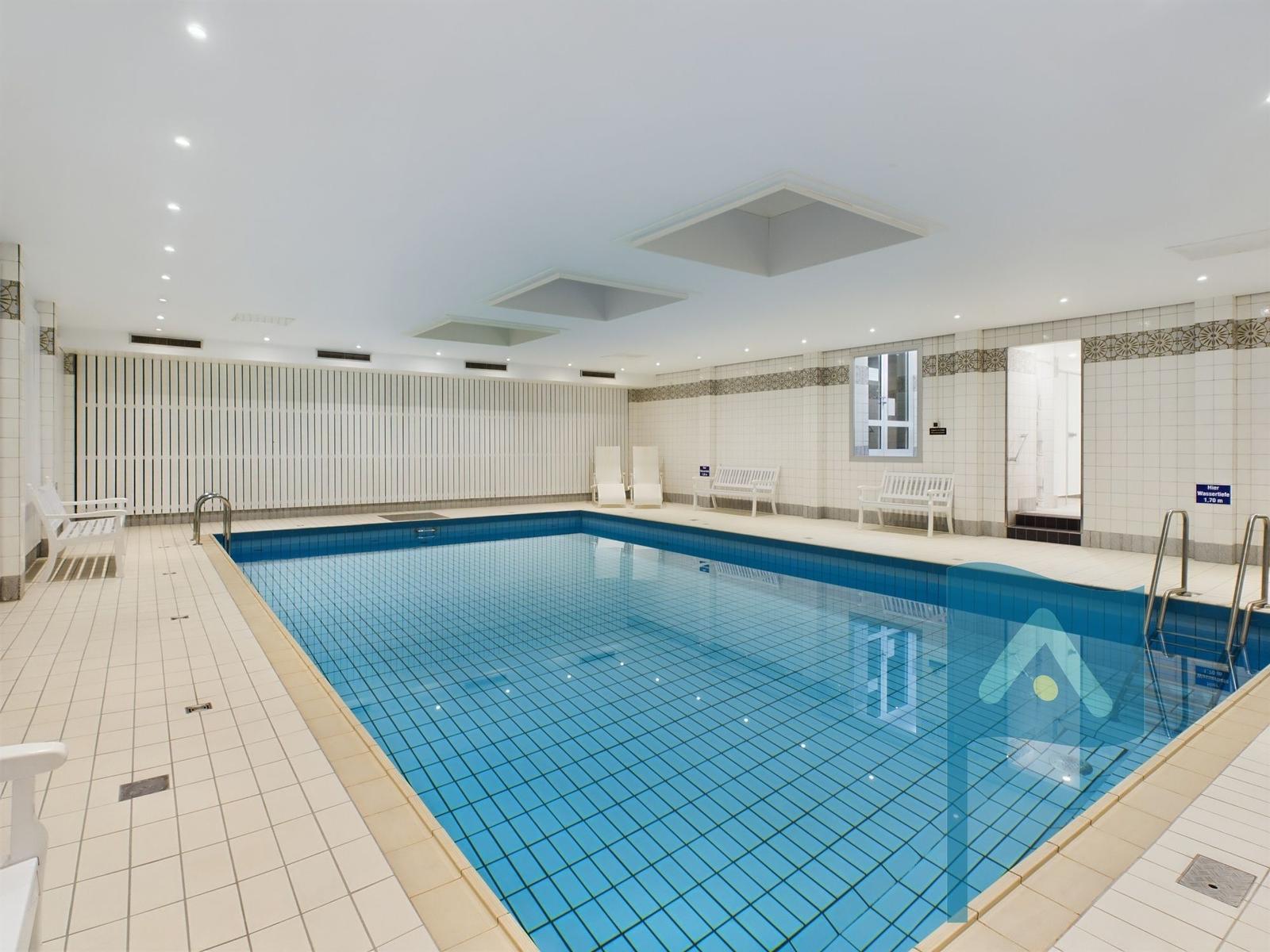 Innenpool mit weißen Bänken und blauen Fliesen. Deckenleuchten und Fenster sorgen für Licht.