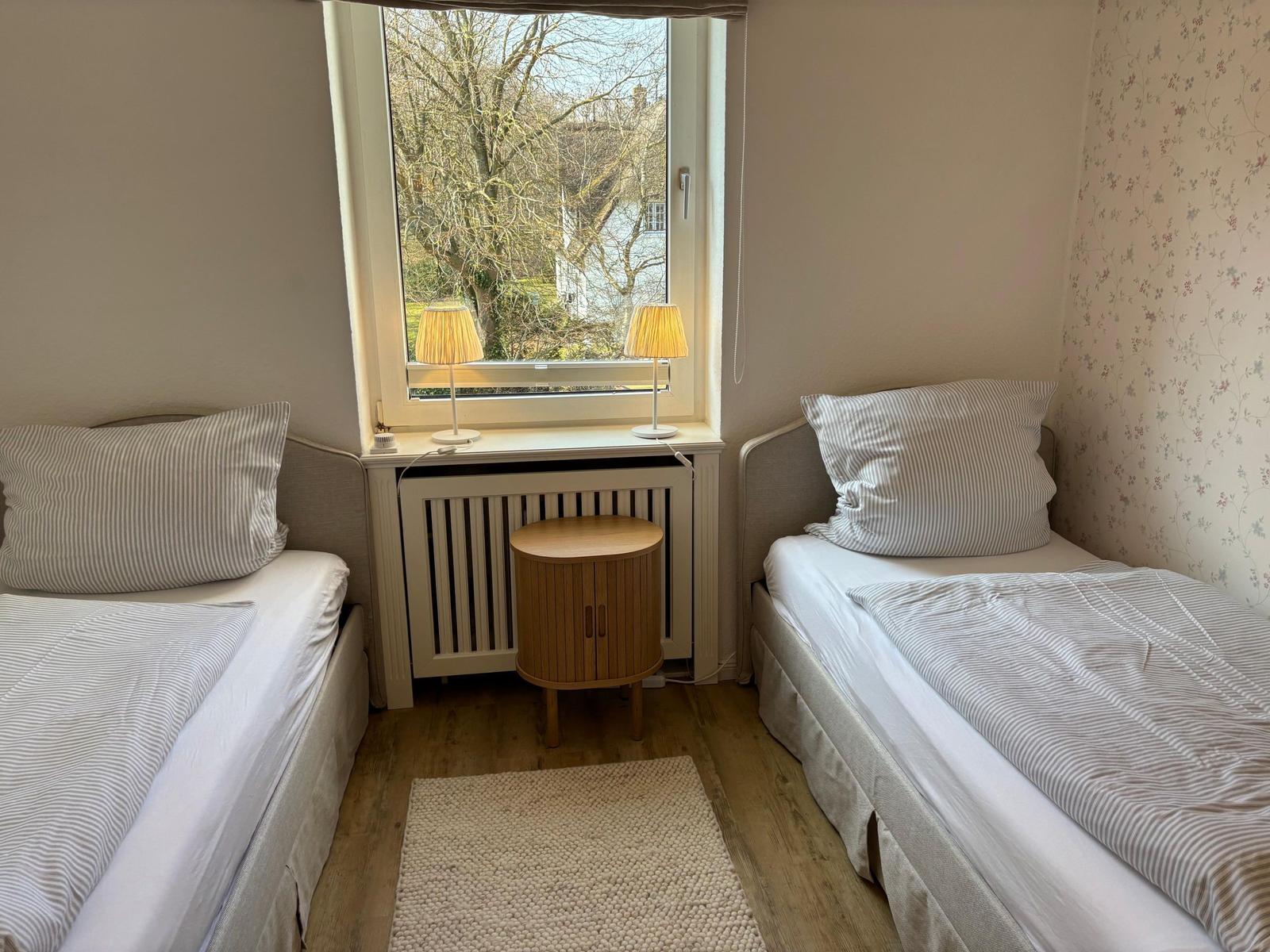Zwei Einzelbetten mit Streifenbetten, Fenster mit Blick auf Bäume, Holztisch und Teppich.