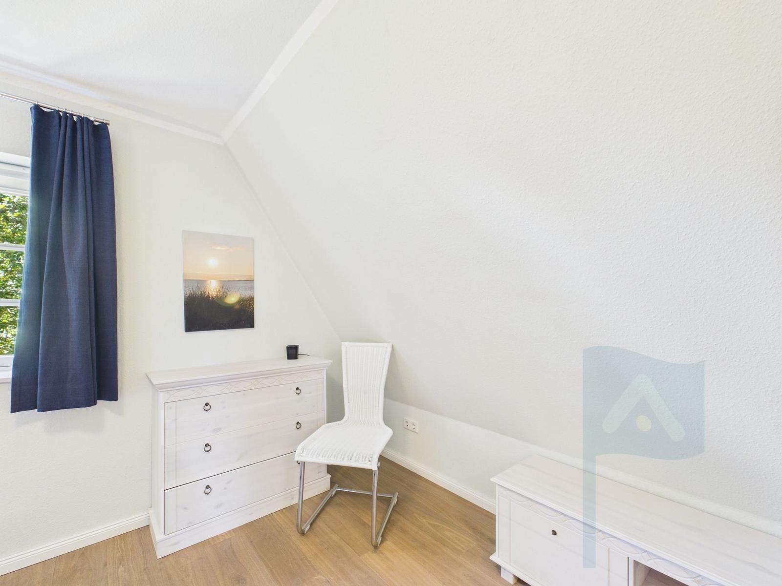 Schlafzimmer mit weißem Schrank, Stuhl und Wandbild. Fenster mit blauen Vorhängen.