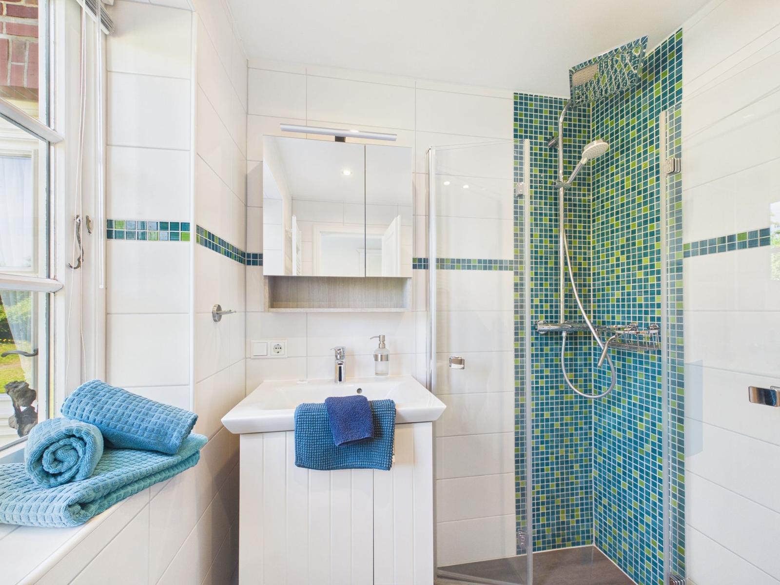 Badezimmer mit Dusche, Waschbecken und Fenster. Mosaikfliesen an der Duschwand.