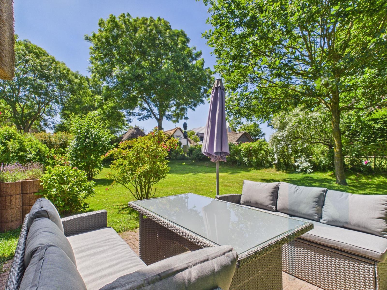 Garten mit Sitzgruppe, Tisch und Schirm. Grünfläche und Bäume im Hintergrund.