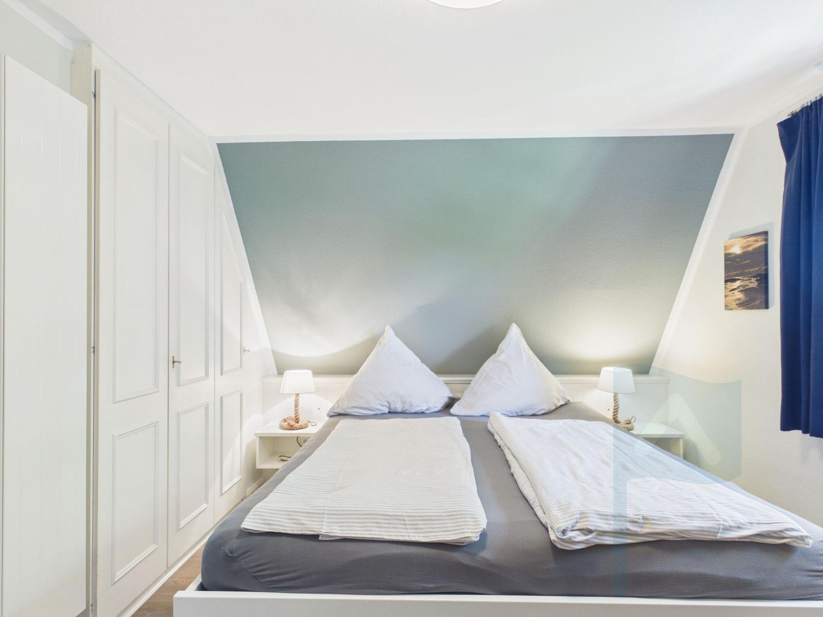 Schlafzimmer mit Doppelbett, Nachttischen und Lampen. Wand in Blautönen. Kleiderkammer und Vorhang.