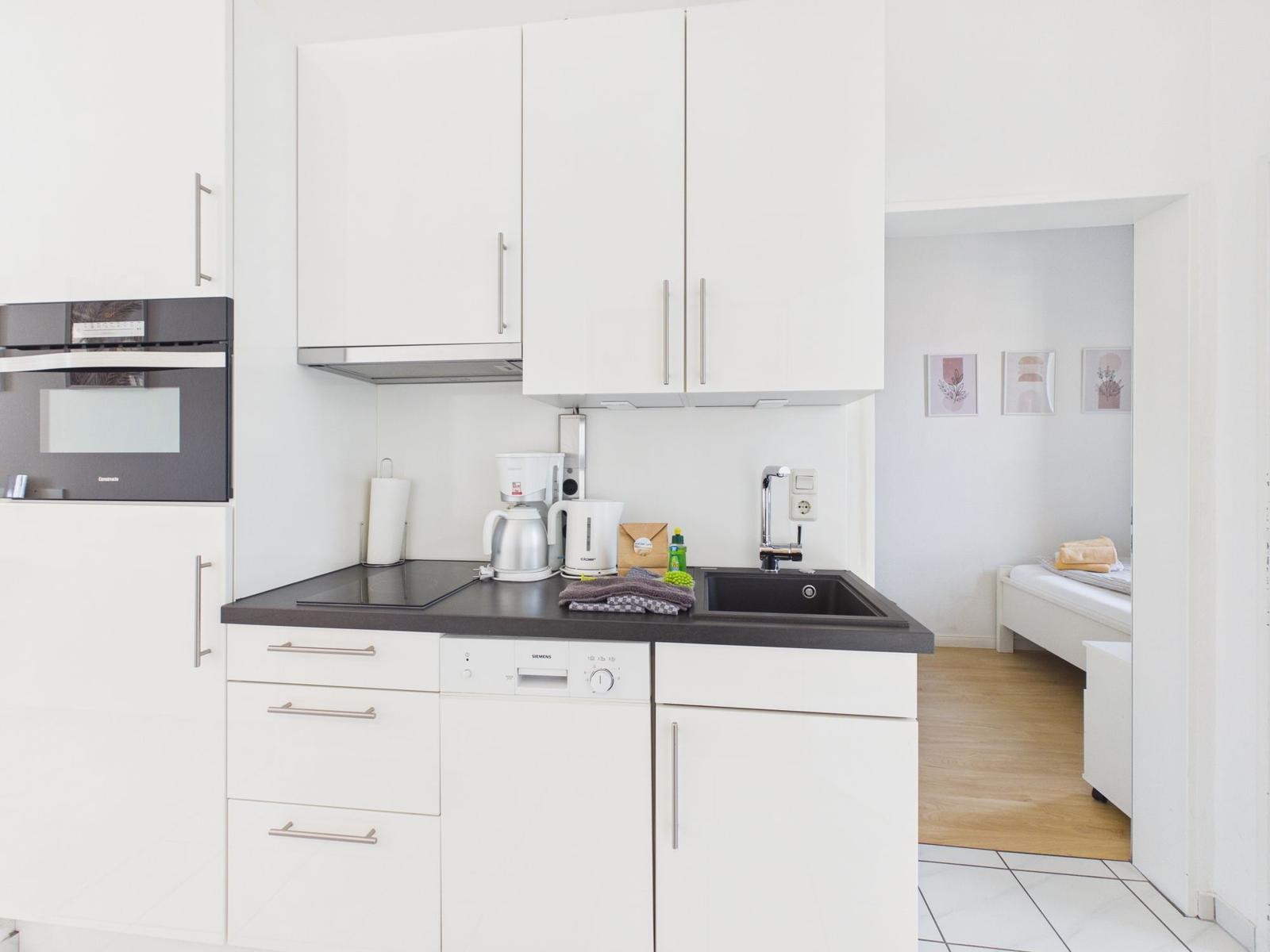 Kitchen with white cabinets, sink, and cooktop. View into bedroom with bed.
