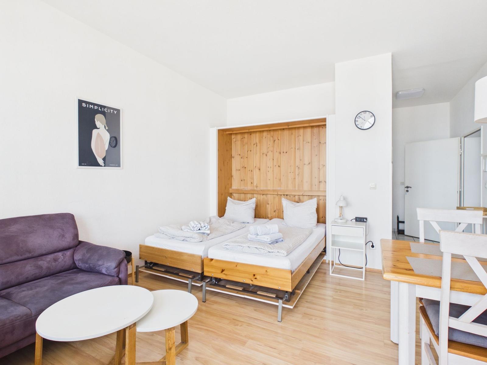 Room with two beds, sofa, table, and wall clock. Wooden surfaces and white walls.