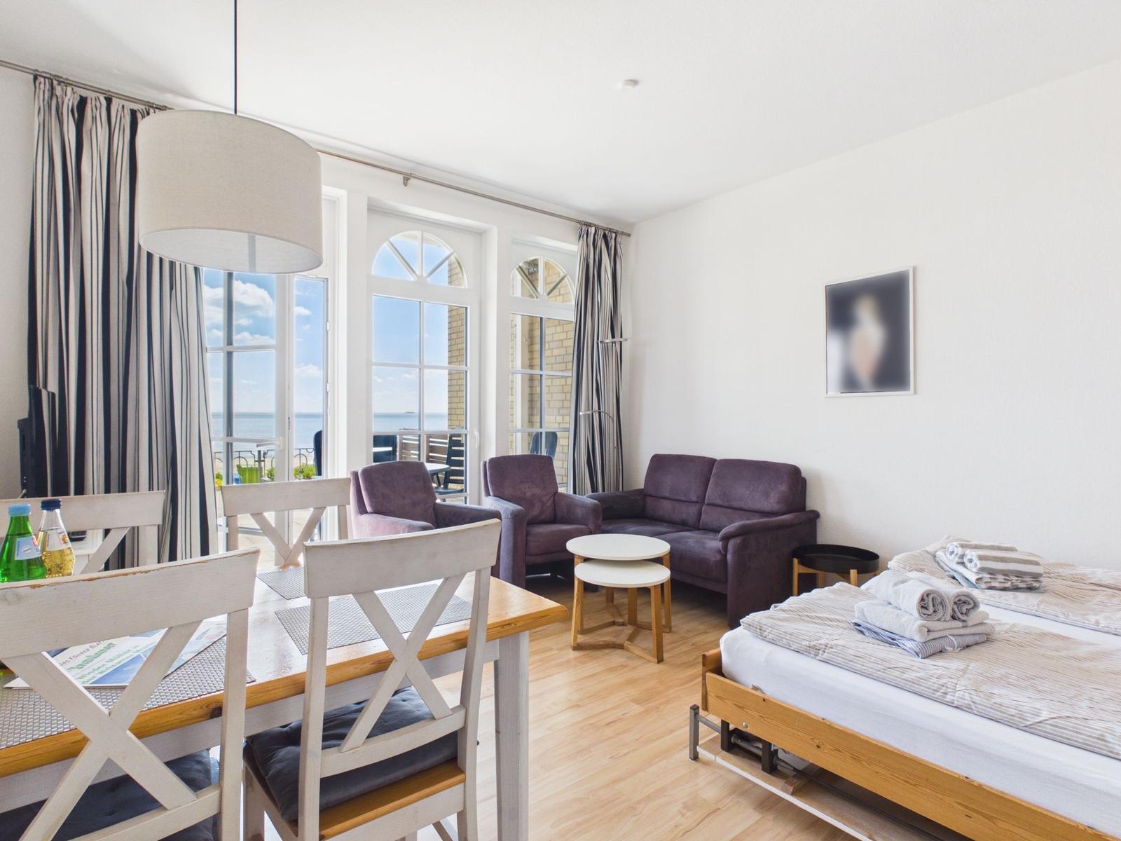 A room with a bed, sofa, table and chairs. Through windows you can see the sea.