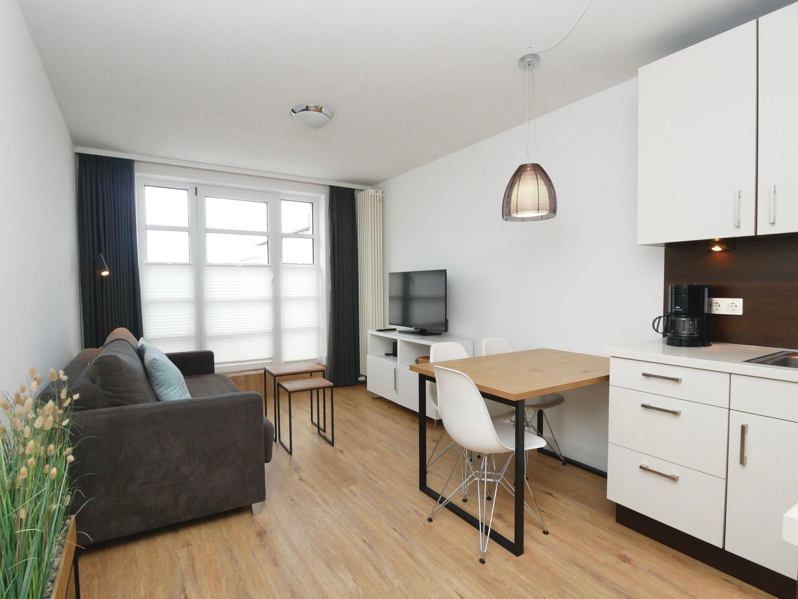 Wohnzimmer mit Sofa, Esstisch und Küchenzeile. Holzboden und große Fenster.