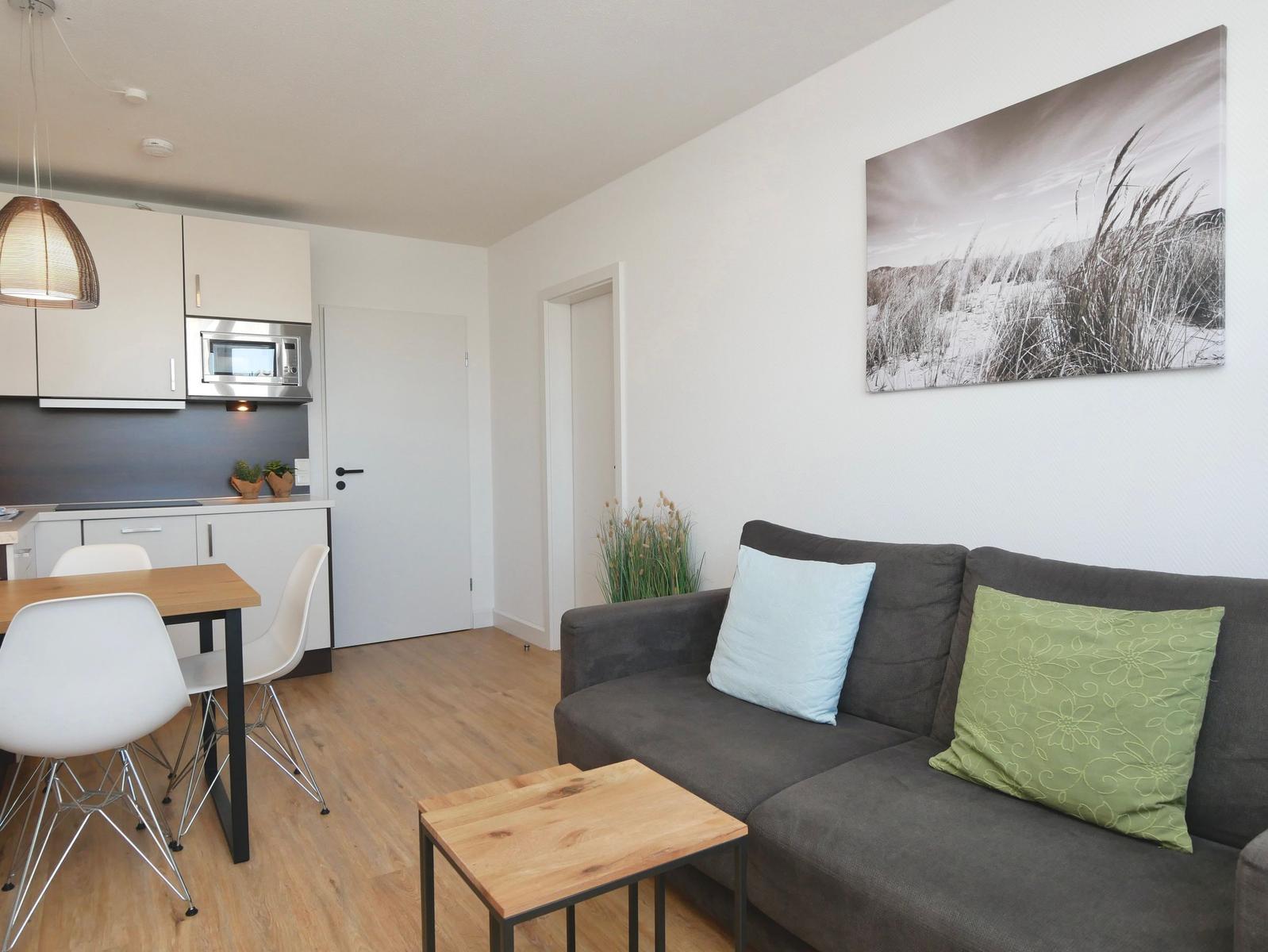 Wohnzimmer mit Sofa, Couchtisch und Küchenbereich. Wandbild zeigt Strandpflanzen.