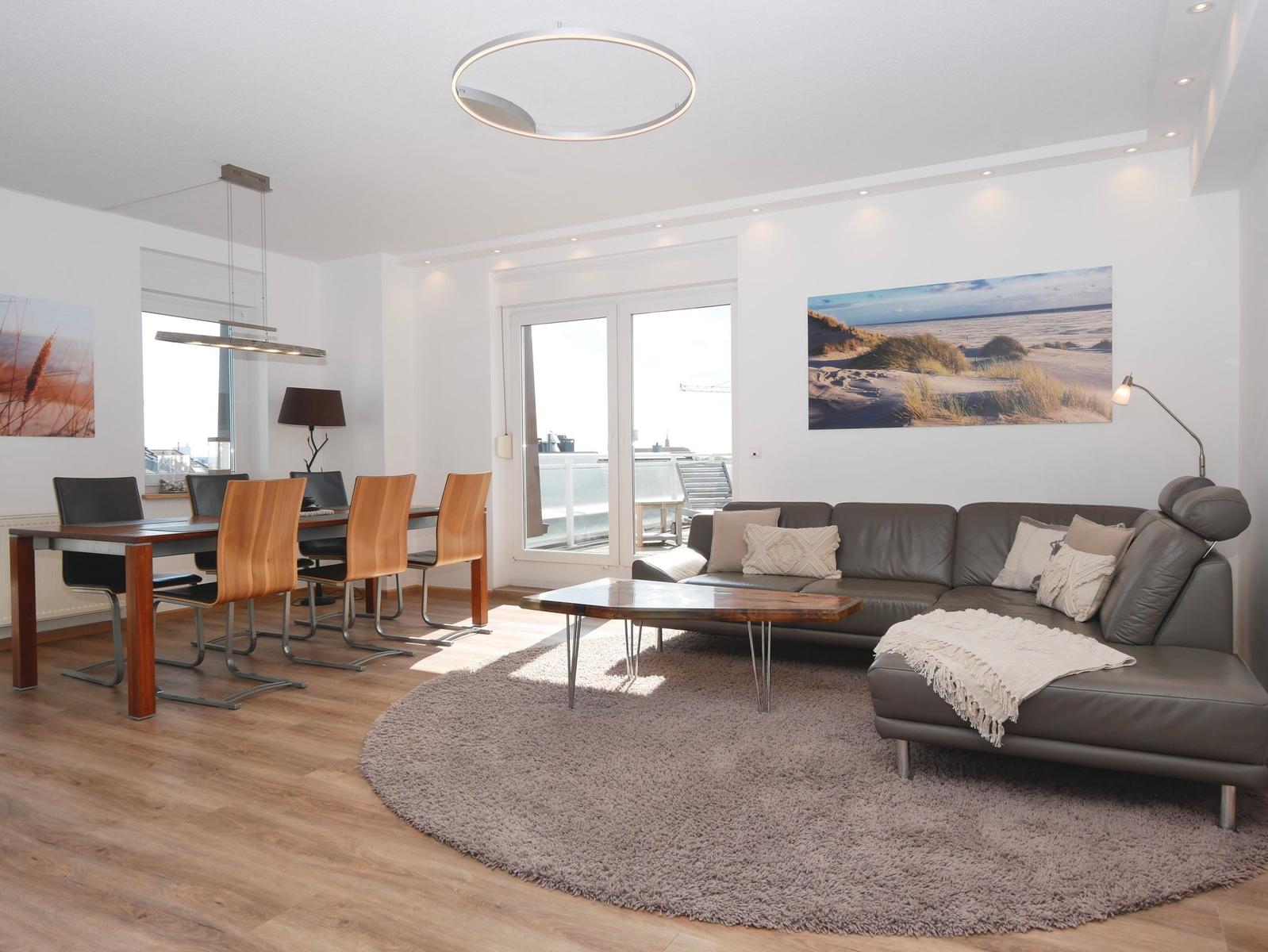 Wohn- und Essbereich mit Sofa, Tisch und Stühlen. Große Fenster und Wandbilder.