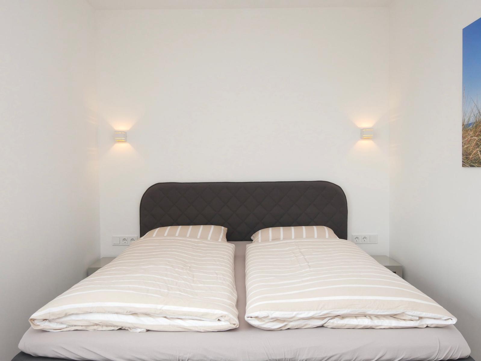 Ein Bett mit gestreiften Bettwäsche und dunklem Kopfteil. Zwei Wandlampen und eine Wanddekoration sind sichtbar.