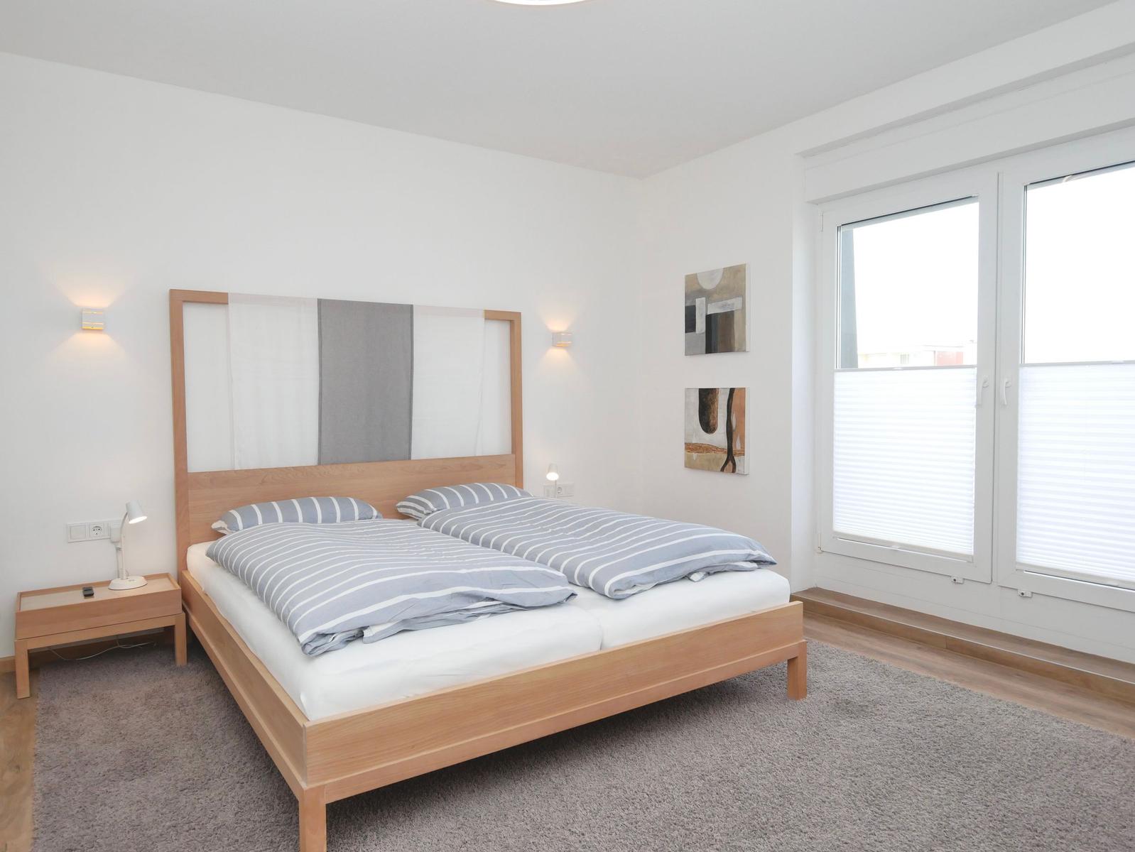 Schlafzimmer mit Doppelbett, Holzgestell, Fenster mit Jalousien und Wandbildern.