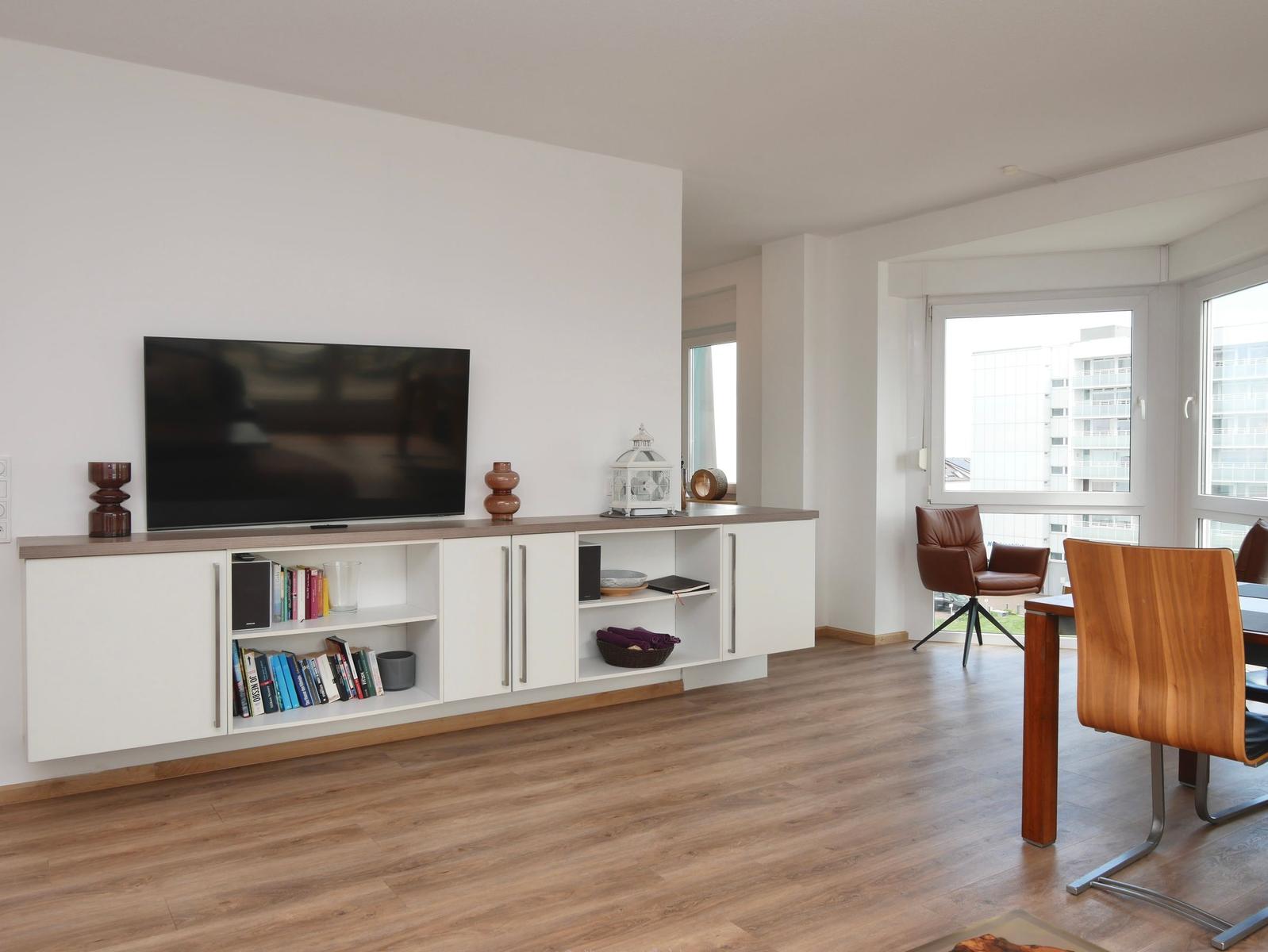 Wohnzimmer mit Fernseher, Bücherregal und Essbereich mit Holzstühlen.