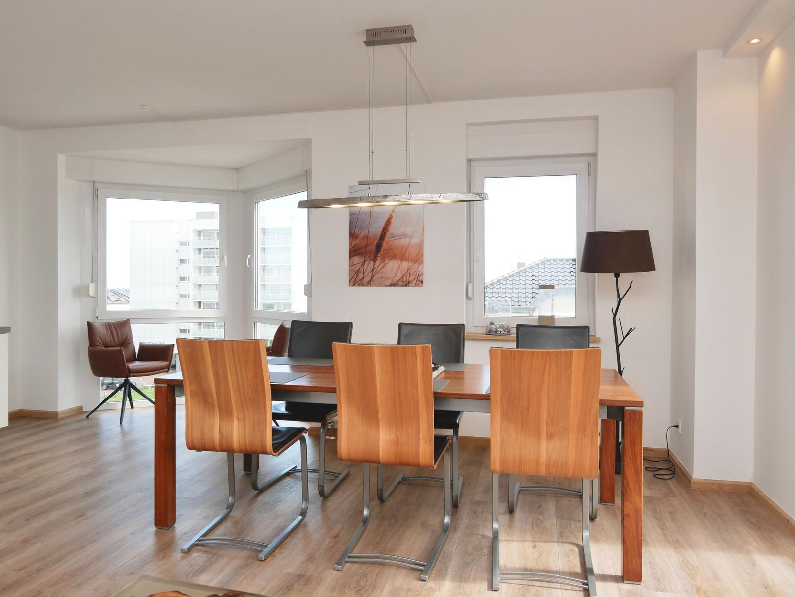 Esszimmer mit Holztisch, Stühlen und Fenstern. Hängelampe und Stehlampe sind sichtbar.