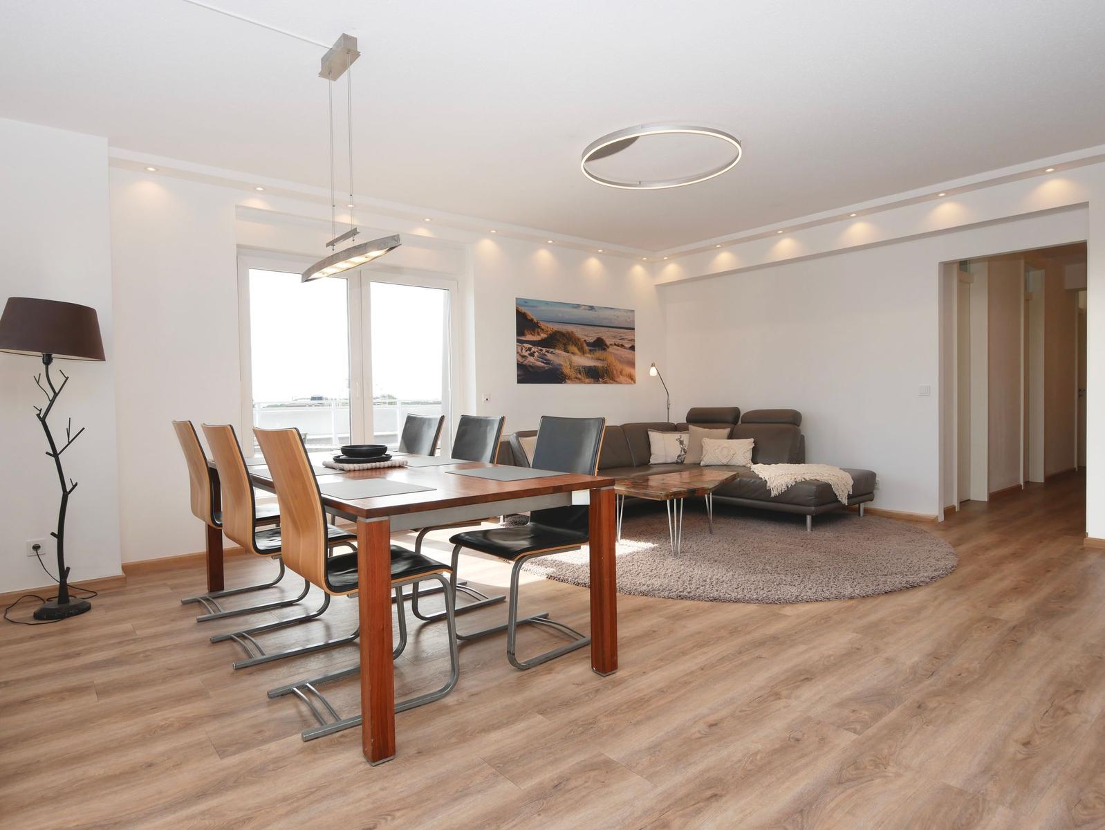 Essbereich mit Tisch und Stühlen. Wohnbereich mit Sofa und Teppich. Holzboden und große Fenster.