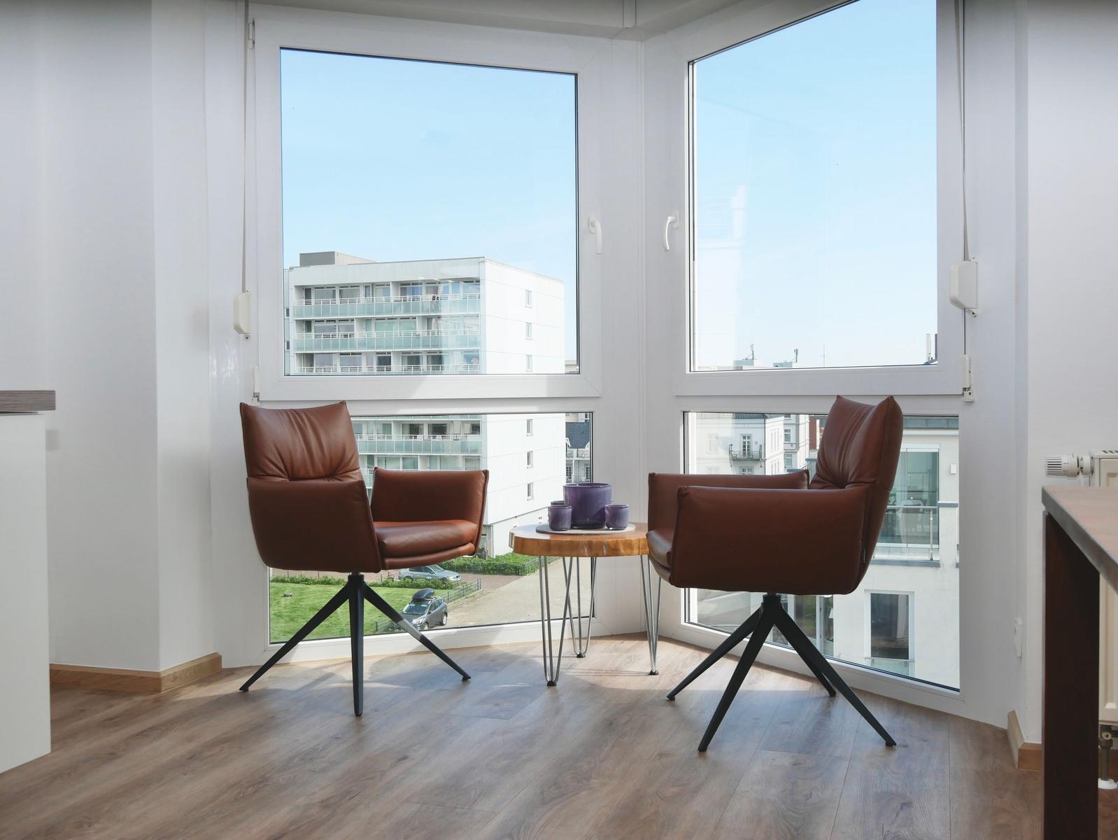 Zwei braune Sessel und ein runder Tisch stehen vor großen Fenstern mit Blick auf Gebäude.