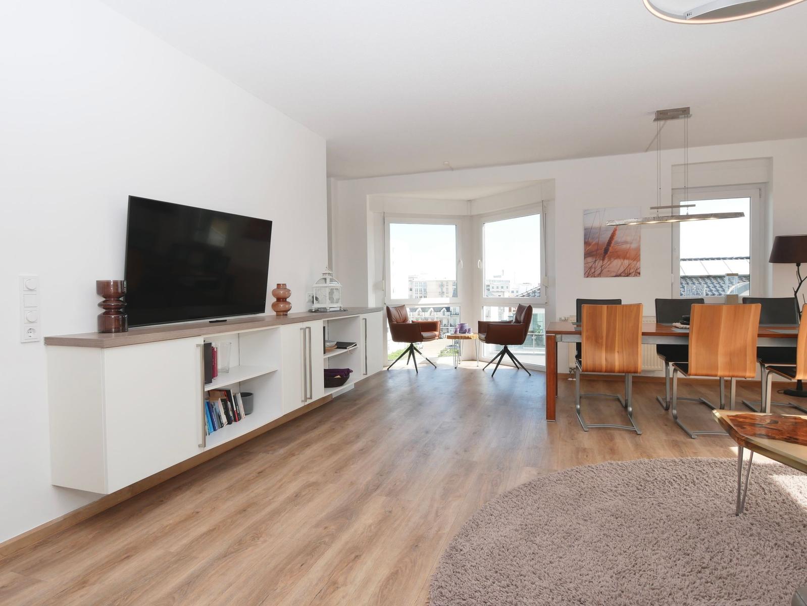Wohn- und Essbereich mit TV, Holzboden und großem Fenster. Möbel sind modern und funktional.