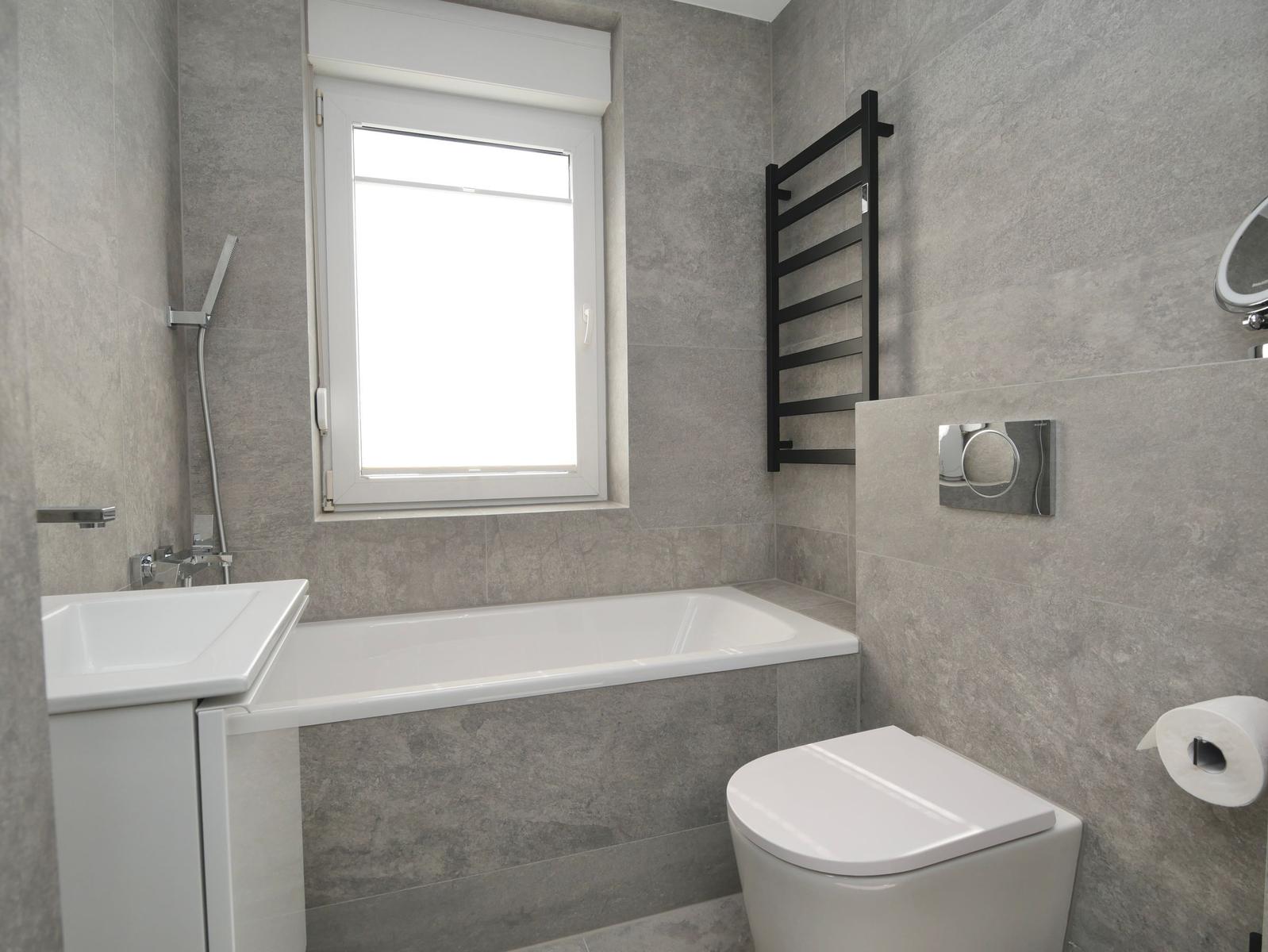 Badezimmer mit Badewanne, Toilette und Waschbecken. Graue Fliesen und Fenster.