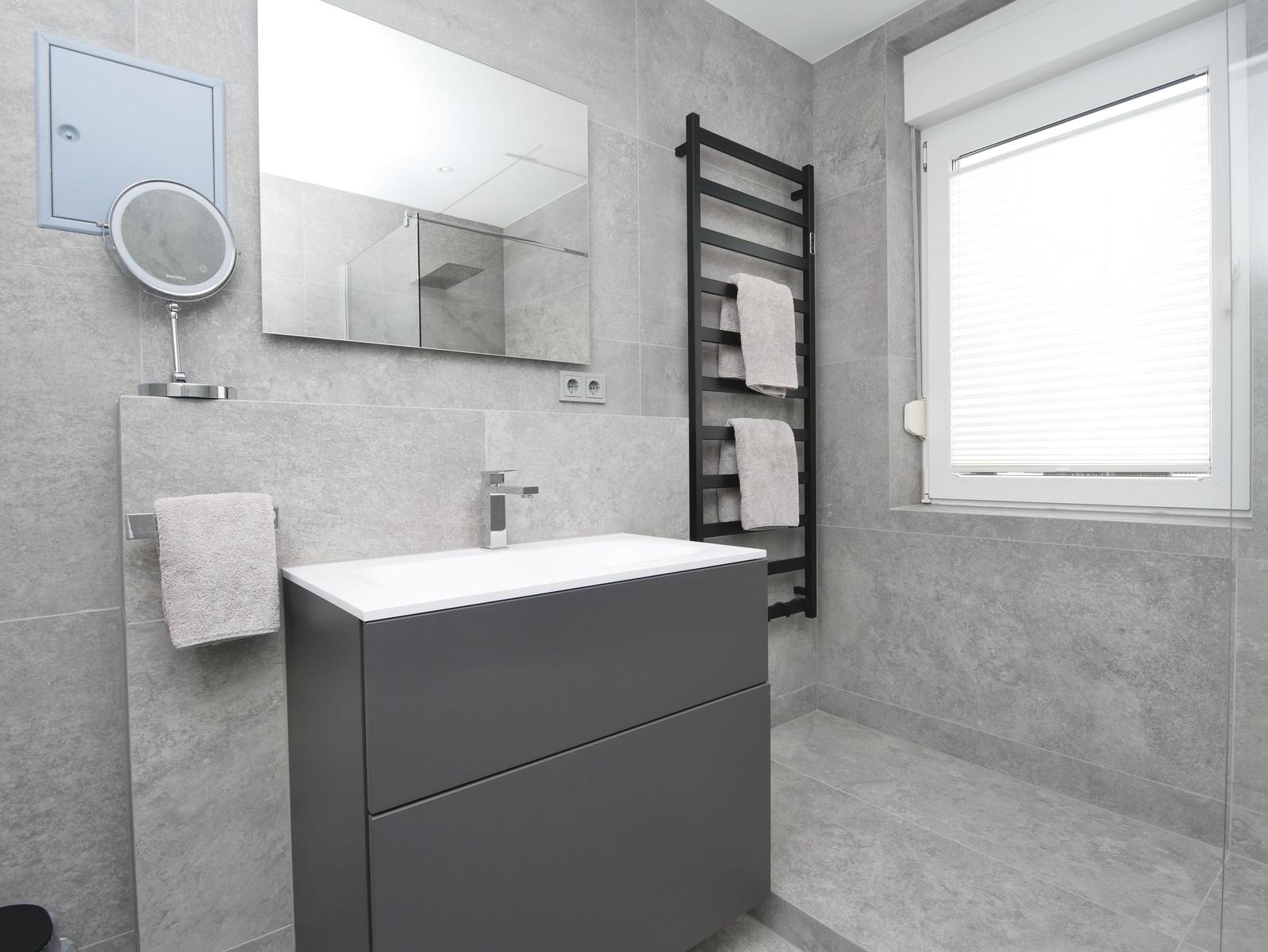 Badezimmer mit Waschbecken, Spiegel und Fenster. Handtuchhalter und Heizkörper sind sichtbar.
