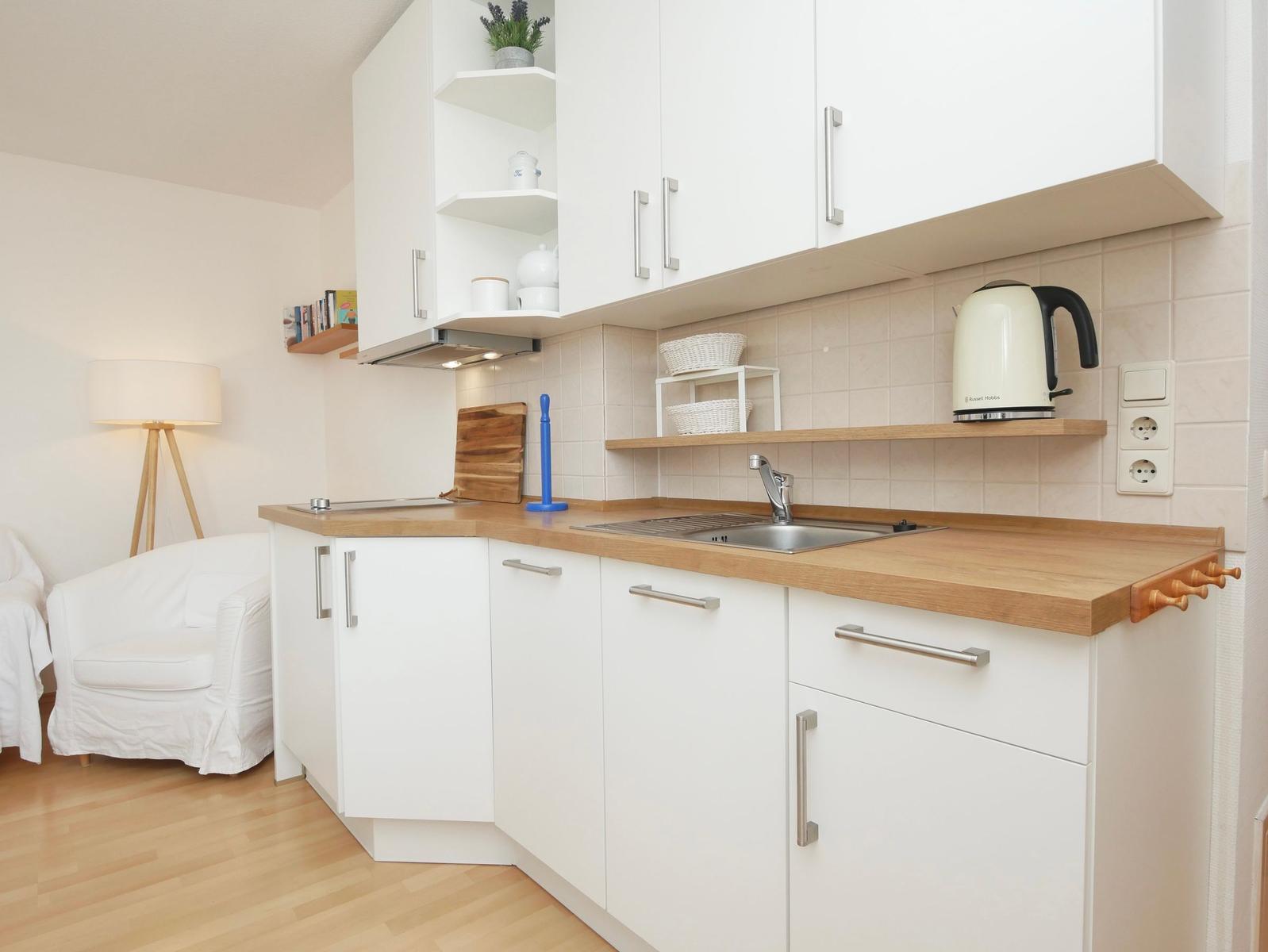 Küche mit weißen Schränken, Holzplatte und Kaffeemaschine. Im Hintergrund Sessel und Stehlampe.