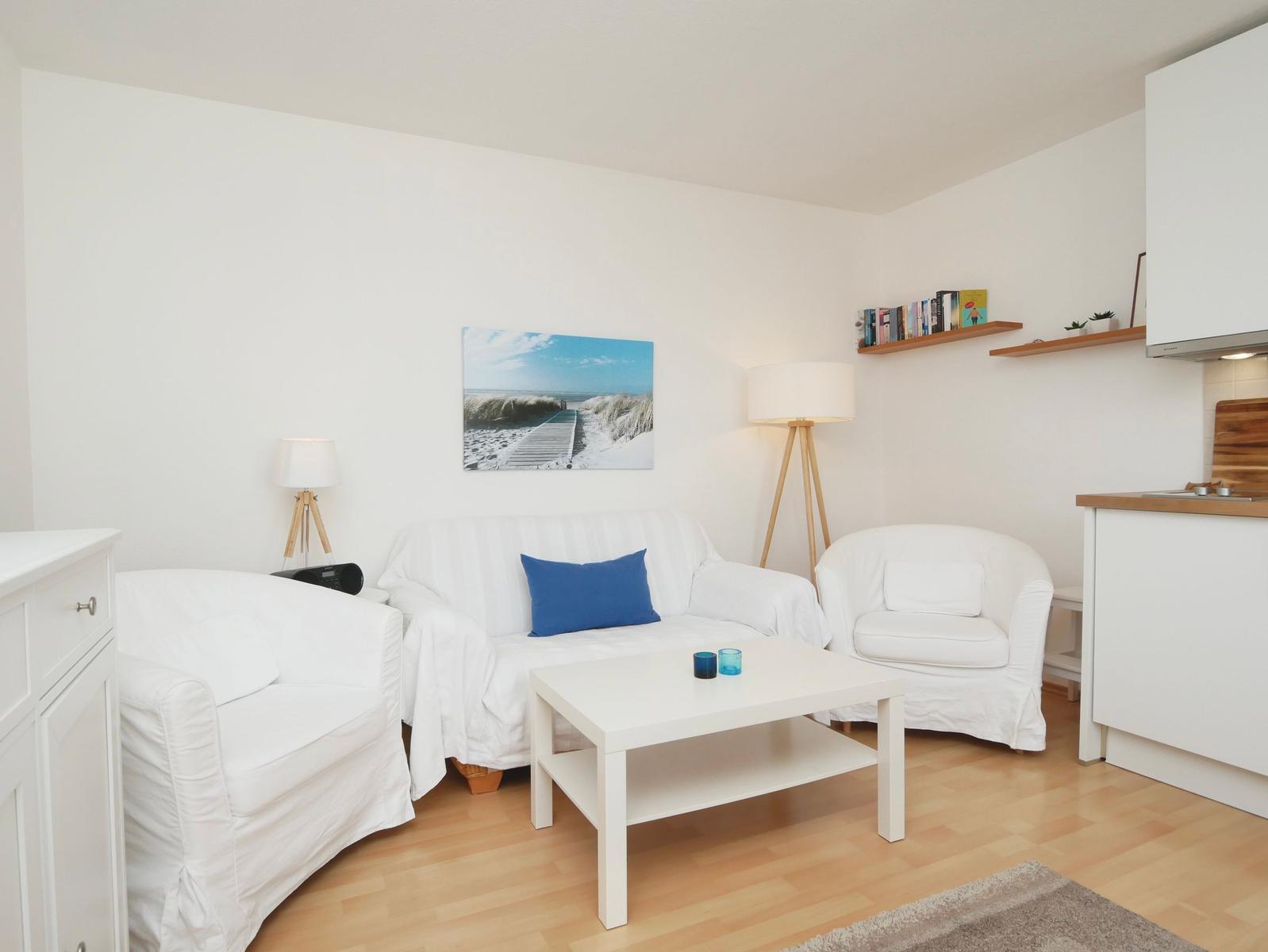 Wohnzimmer mit weißem Sofa, zwei Sesseln und Holzboden. Wandbild und Regale sind sichtbar.