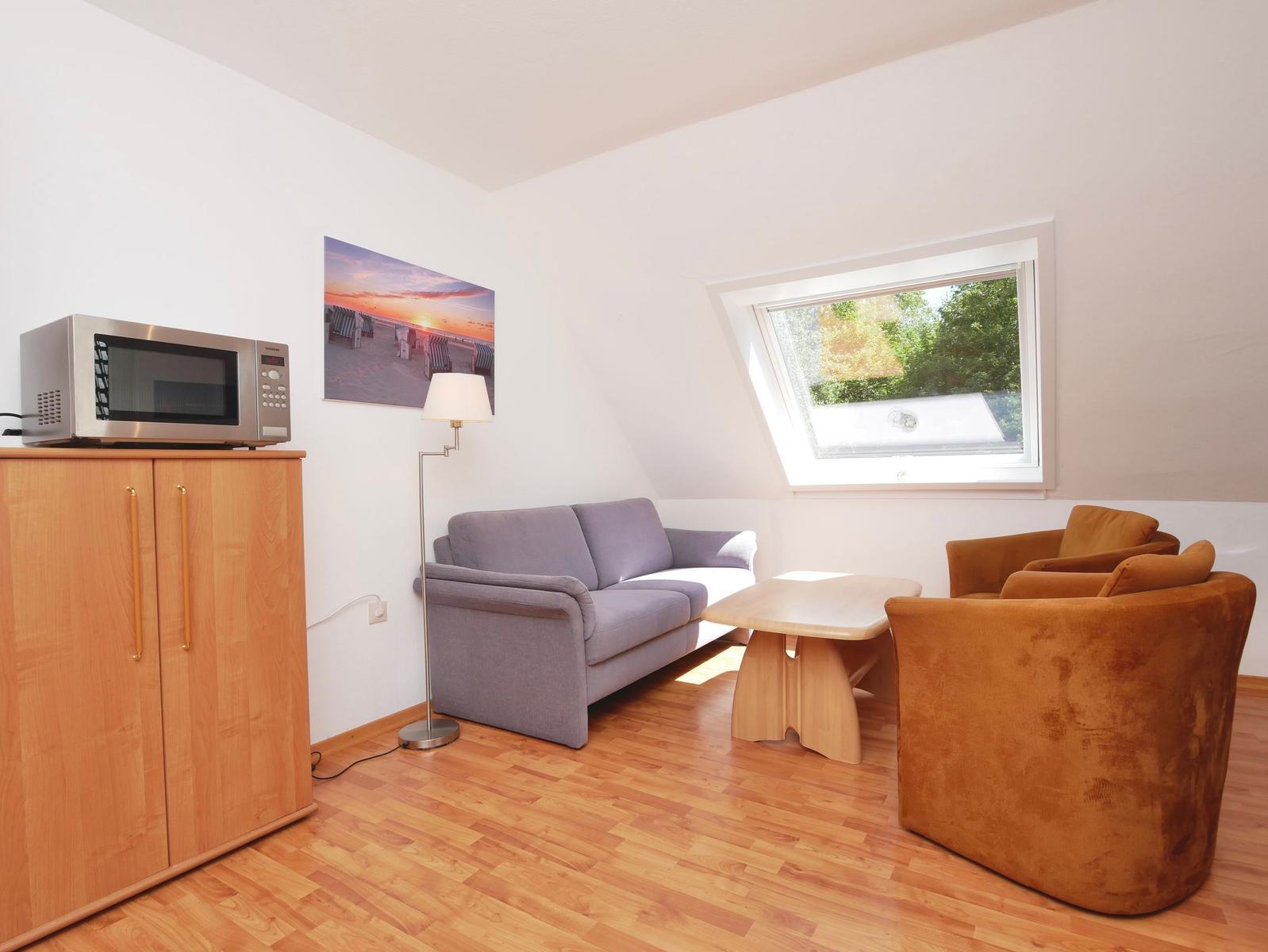 Wohnzimmer mit Sofa, zwei Sesseln, Tisch und Mikrowelle. Dachfenster mit Blick auf Grün.