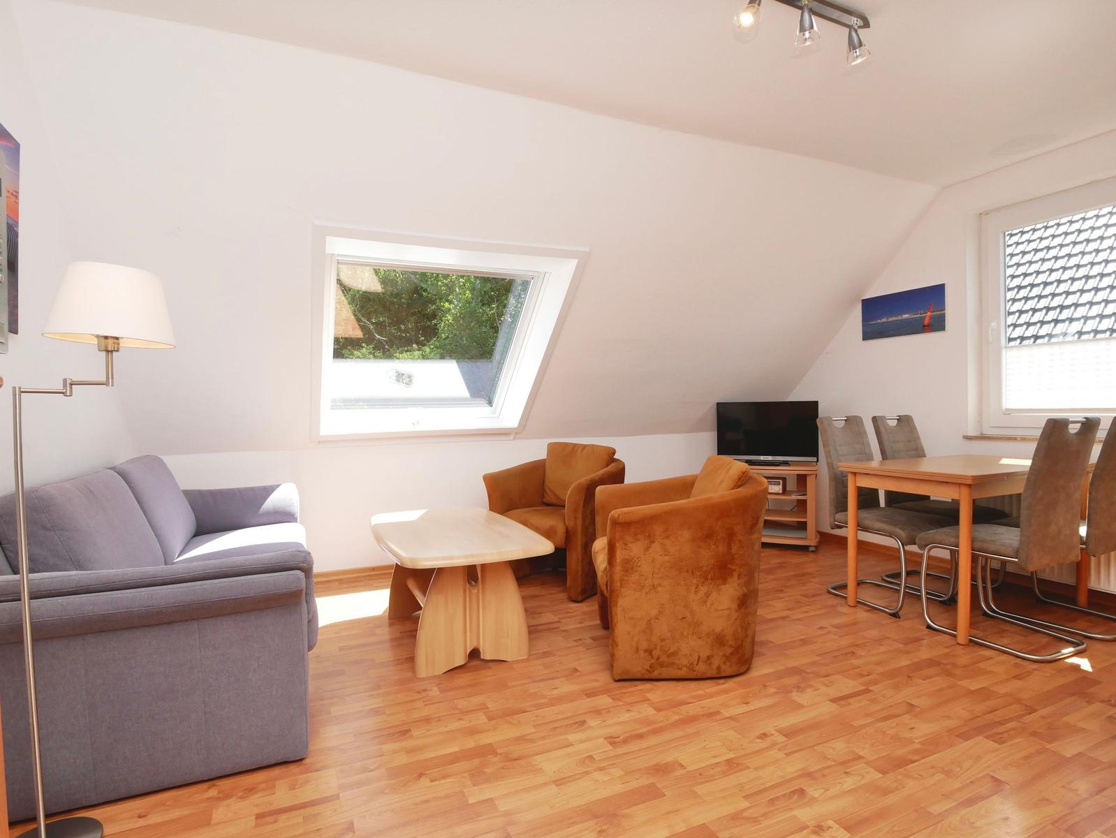 Wohn- und Essbereich mit Sofa, Sesseln, Tisch und TV. Holzboden und Dachschrägen.