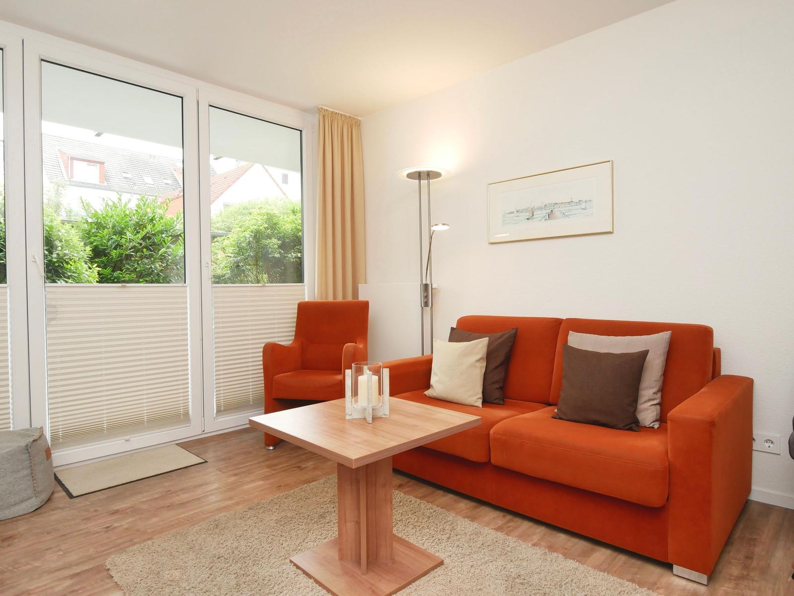 Ein Wohnzimmer mit orangefarbenem Sofa, Sessel und Holztisch. Große Fenster mit Blick auf Grünflächen.