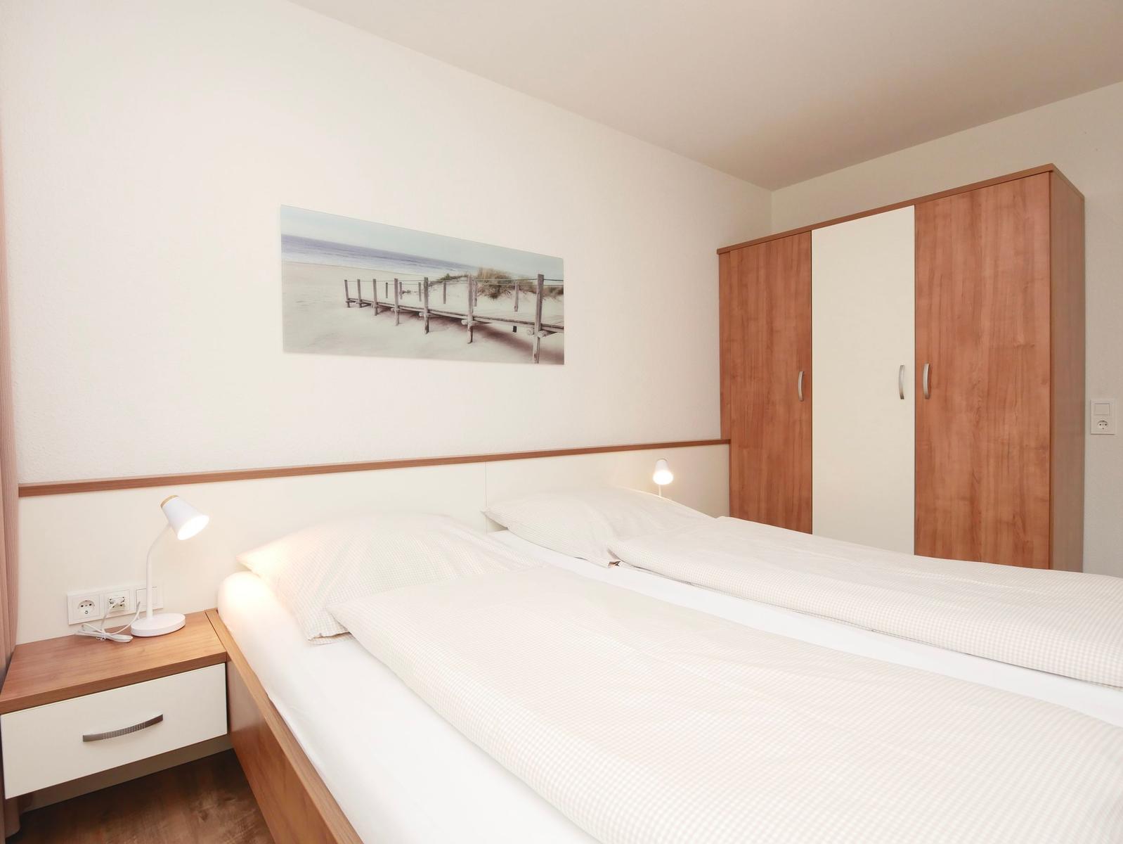 Schlafzimmer mit Doppelbett, Nachttisch und Kleiderschrank. Wandbild zeigt Strand.