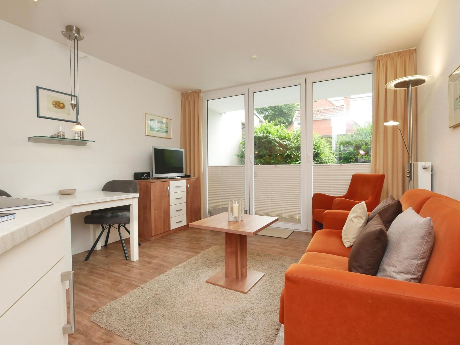 Wohnzimmer mit orangefarbenem Sofa, Tisch und TV. Durchgang zur Terrasse mit Blick auf Grün.
